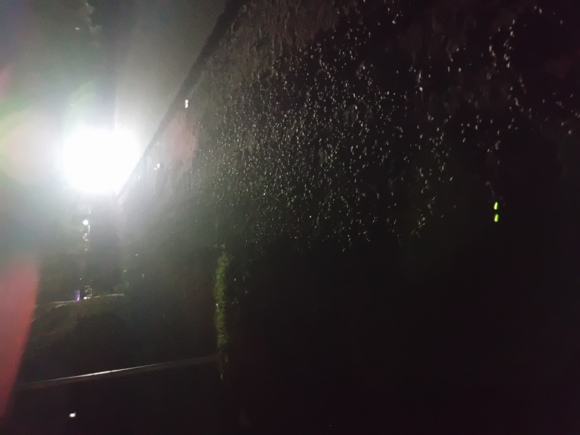 An outdoor nighttime scene shows a dark, unpaved road. The road is covered with small stones. To the left of the road is a patch of dark grass and a vertical pole. In the distance, a bright, white light illuminates the road and the surrounding area, obscuring details. There are two small, green lights visible on the road in the foreground, possibly reflective markers or the eyes of an animal. The sky is dark and appears to be over a rural or undeveloped area.