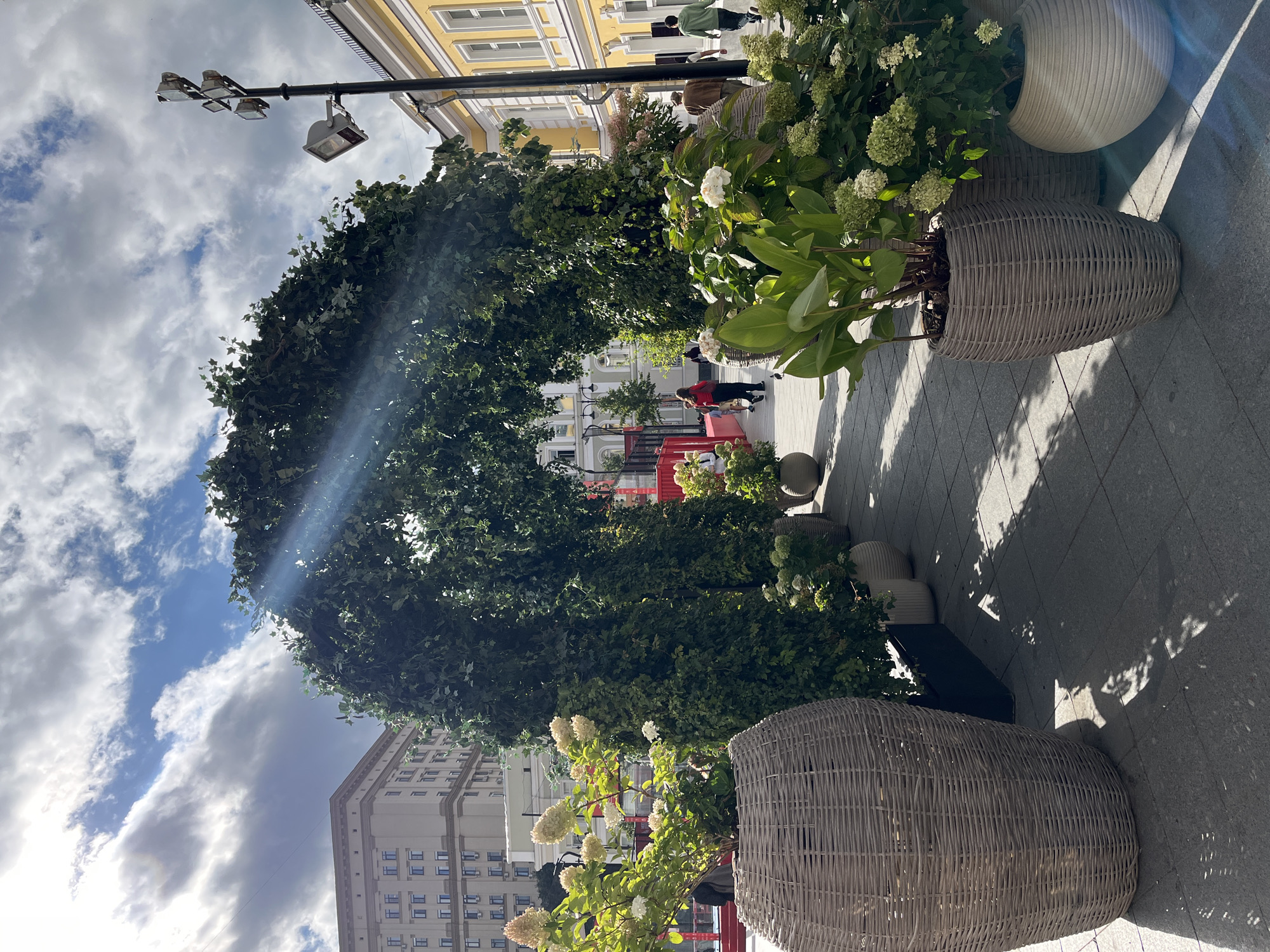 This is an outdoor photograph taken in a city. In the foreground, there are several large planters made of woven material, containing lush green plants and white flowers. These planters line a paved walkway. Dominating the center of the image is a large, arched structure covered in dense green foliage, forming an overhead canopy. Sunlight streams through the leaves, creating bright highlights and shadows on the ground. In the background, there are buildings of varying architectural styles, one appearing to be a multi-story apartment building with numerous windows, and another a lighter-colored building with yellow accents and many windows. Several people are visible walking along the path, some interacting with the environment or each other, while others are simply passing through. A street lamp with multiple light fixtures is visible on the right side of the image. The sky above is partly cloudy with visible sun rays piercing through. The overall scene suggests a public urban space, possibly a pedestrian street or a plaza, adorned with decorative greenery.