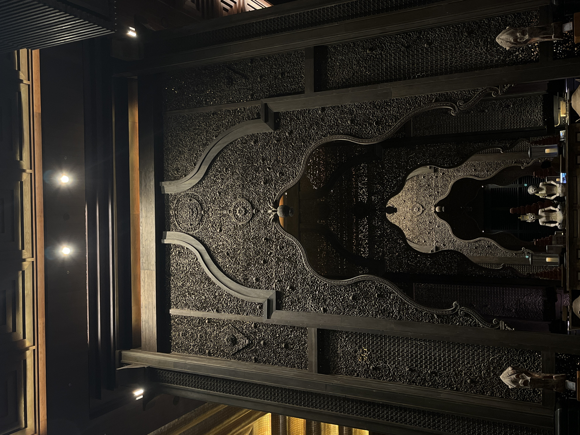 This image depicts an interior scene with ornate, dark wooden carvings and architectural elements. The focal point is a large, intricately carved wooden screen featuring multiple arched openings, reminiscent of Islamic or Indian architectural styles. The screen is heavily textured with detailed patterns, possibly depicting floral motifs or geometric designs.
In the foreground, on a wooden surface, are several decorative objects and figures. To the left, there is a carved wooden statue of a head or animal. In the center, there are at least two seated Buddha-like statues with elaborate headdresses. A candle in a glass holder is visible to the right of the statues, along with other indiscernible items on the surface. The background features more carved wooden structures, including pillars and overhead beams, with subtle lighting that highlights the textures and depths of the carvings. The lighting appears to be artificial, with spotlights integrated into the ceiling structure.
The environment suggests a formal, possibly religious or cultural, interior space. The scale of the carved screen and its intricate detail indicate significant craftsmanship and expense. The overall atmosphere is one of grandeur and solemnity.
While the image itself does not provide explicit geographical indicators, if this scene is indeed located in Kuta Selatan, Indonesia, it would likely be within a high-end resort, a cultural center, or a significant religious building that incorporates such elaborate architectural and decorative elements. Kuta Selatan is known for its luxurious accommodations and tourist attractions, which often feature distinctive architectural styles that blend local influences with international design.