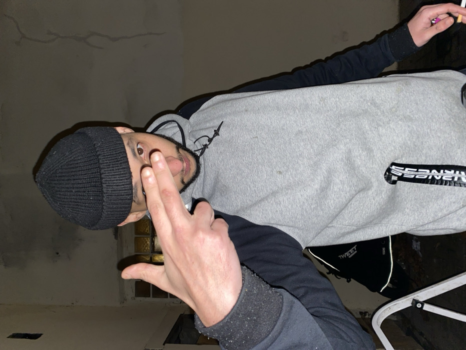 A man wearing a black beanie and a gray and black hoodie makes a hand gesture with two fingers raised. He has a beard and mustache. In his other hand, he holds a cigarette. He appears to be indoors with a window behind him. The wall behind him is beige with a few cracks. He is standing near a stepladder.
