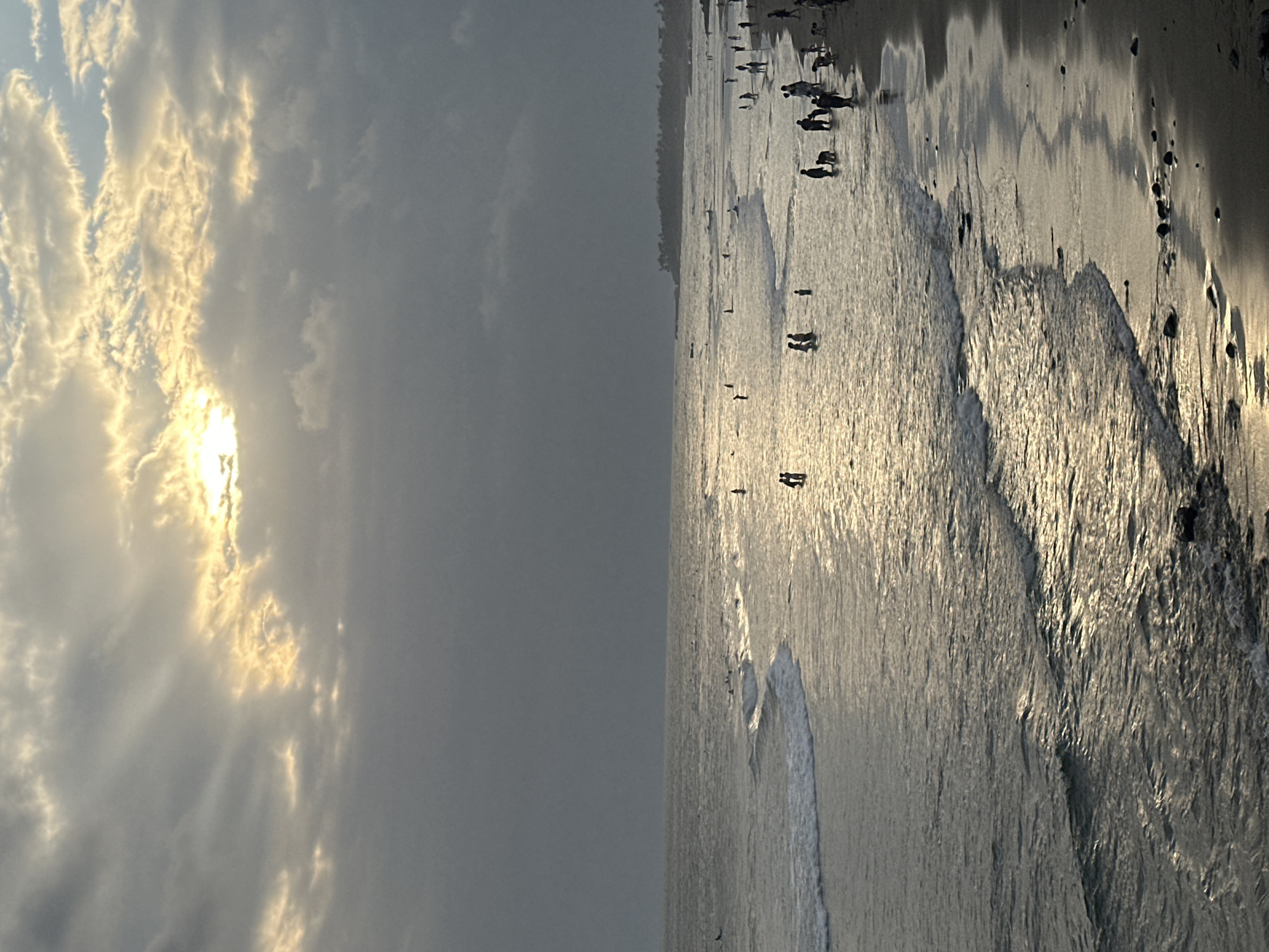 The image displays a serene beach scene at sunset in North Kuta, Indonesia. The sky is partially cloudy, with the sun casting a warm, golden glow through the clouds, creating a dramatic effect with sunbeams radiating outwards. The ocean is calm, with gentle waves lapping at the shore. The wet sand reflects the sky and sunlight, creating a shimmering surface. Several people are visible on the beach, either walking along the shoreline or standing near the water's edge. Their figures are silhouetted against the bright sand and sea, suggesting a peaceful and leisurely atmosphere. The overall mood is tranquil, evoking a sense of calm and natural beauty as the day draws to a close. There is no visible text in the image.