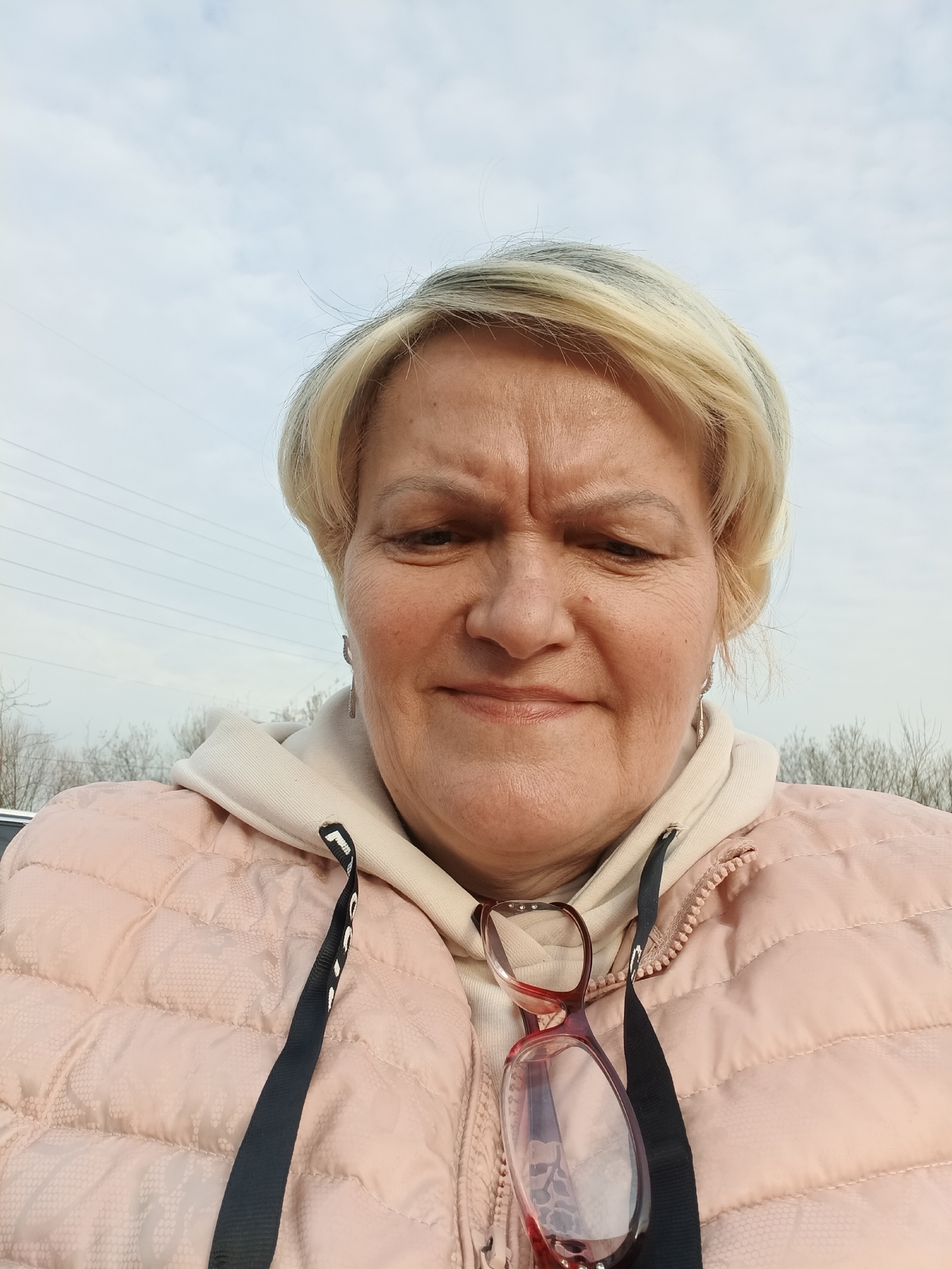 A female subject, appearing middle-aged with short blonde hair, is depicted in a medium-close-up, eye-level outdoor portrait. Her expression is neutral with a slightly furrowed brow, and she looks directly at the camera. She wears a light pink, textured, quilted jacket over a light-colored hooded sweatshirt. A pair of reddish-brown framed glasses is suspended around her neck by a black lanyard and rests on her chest. Small silver-colored earrings are visible. The background features an overcast sky with light clouds. In the mid-ground, multiple parallel power lines stretch horizontally across the left side of the frame. A faint line of bare deciduous trees or shrubs is visible along the distant horizon. The visual evidence does not provide specific markers to confirm a precise location within Odintsovo, Russia; however, the environment is consistent with an open outdoor area on the periphery of a developed zone.