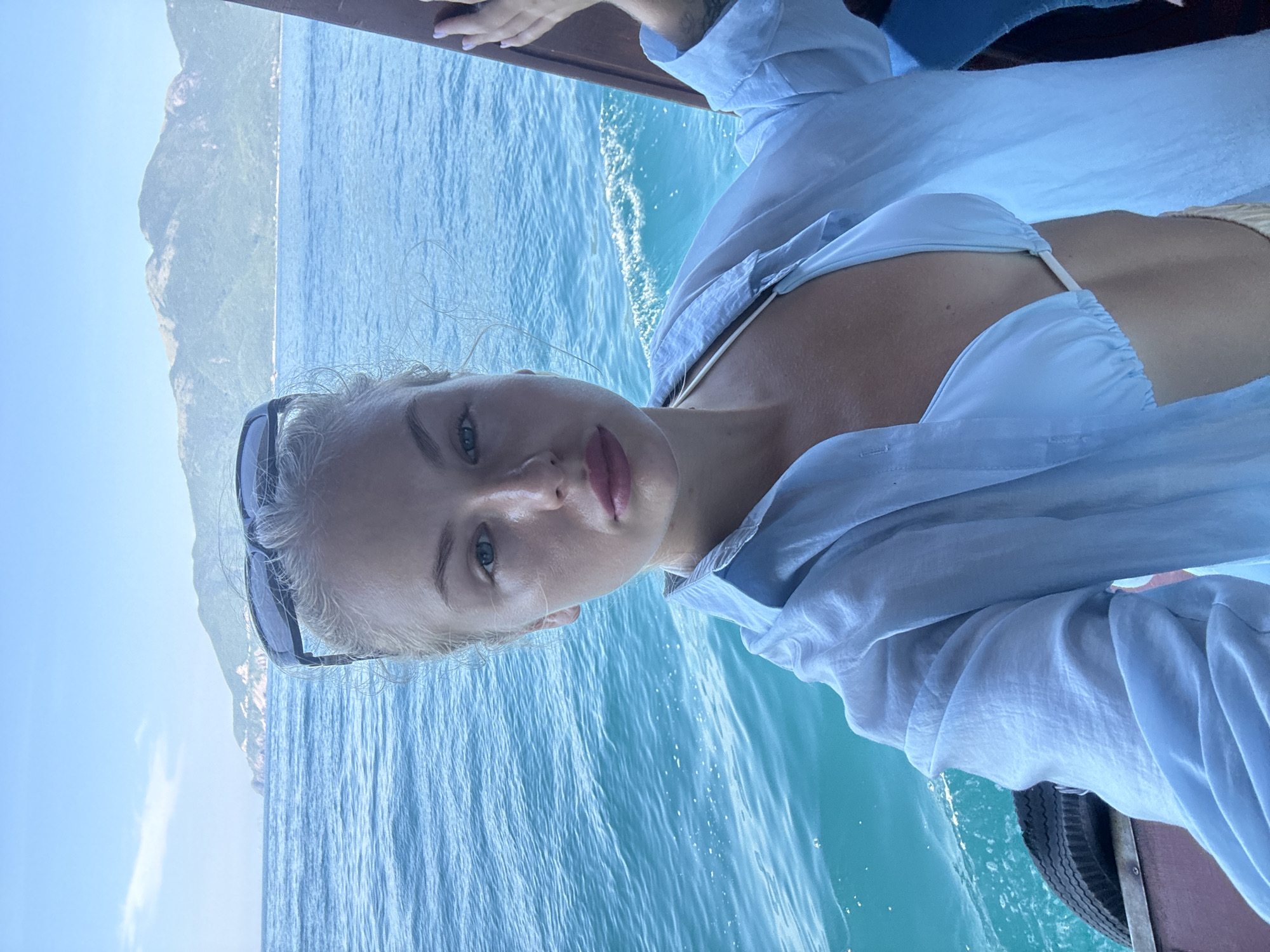 A woman with blonde hair pulled back and sunglasses perched on her head takes a selfie on a boat in Nha Trang, Vietnam. She is wearing a light blue bikini top and an unbuttoned light blue shirt. The ocean is blue and choppy, and in the background, lush green mountains rise from the coastline under a clear sky. She is leaning against the side of the boat, her right hand resting on a wooden railing. A spare tire is visible on the boat's deck.