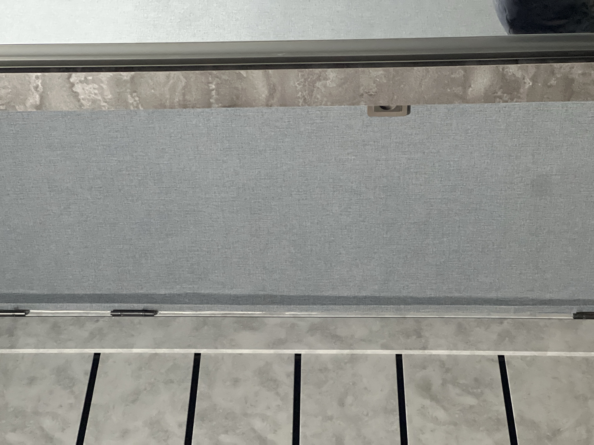 The image shows an indoor scene with a gray textured surface dominating the central view. Above it is a marble-like shelf, possibly a countertop, with a gray trim. Above the shelf, a lighter gray surface is visible, possibly a wall. Below the textured surface is a row of light gray tiles with dark vertical lines separating them. The scene could be part of a bathroom or kitchen, given the materials and layout. The location is unknown but requested to be Yerevan, Armenia. No people or interactions are visible in the image.