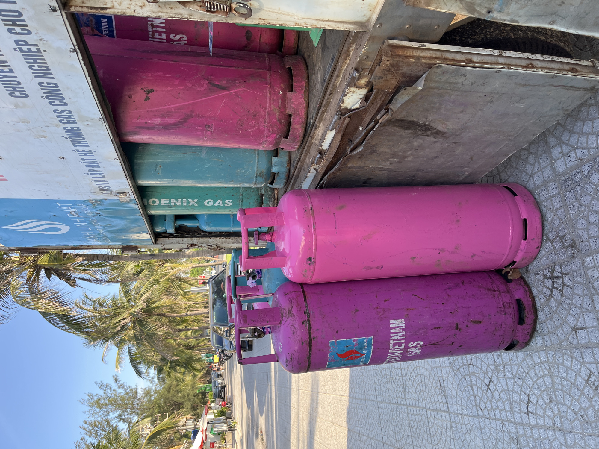The image shows stacked, cylindrical propane gas tanks, some painted pink and purple, labeled "PETROVIETNAM GAS" and "PHOENIX GAS". They are in a truck bed with rusted metal. A sign with Vietnamese text is partially visible. The foreground has patterned pavement. In the background, palm trees and buildings indicate a coastal location, consistent with Đà Nẵng, Vietnam. Vehicles and other street-side elements are discernible in the distant background.