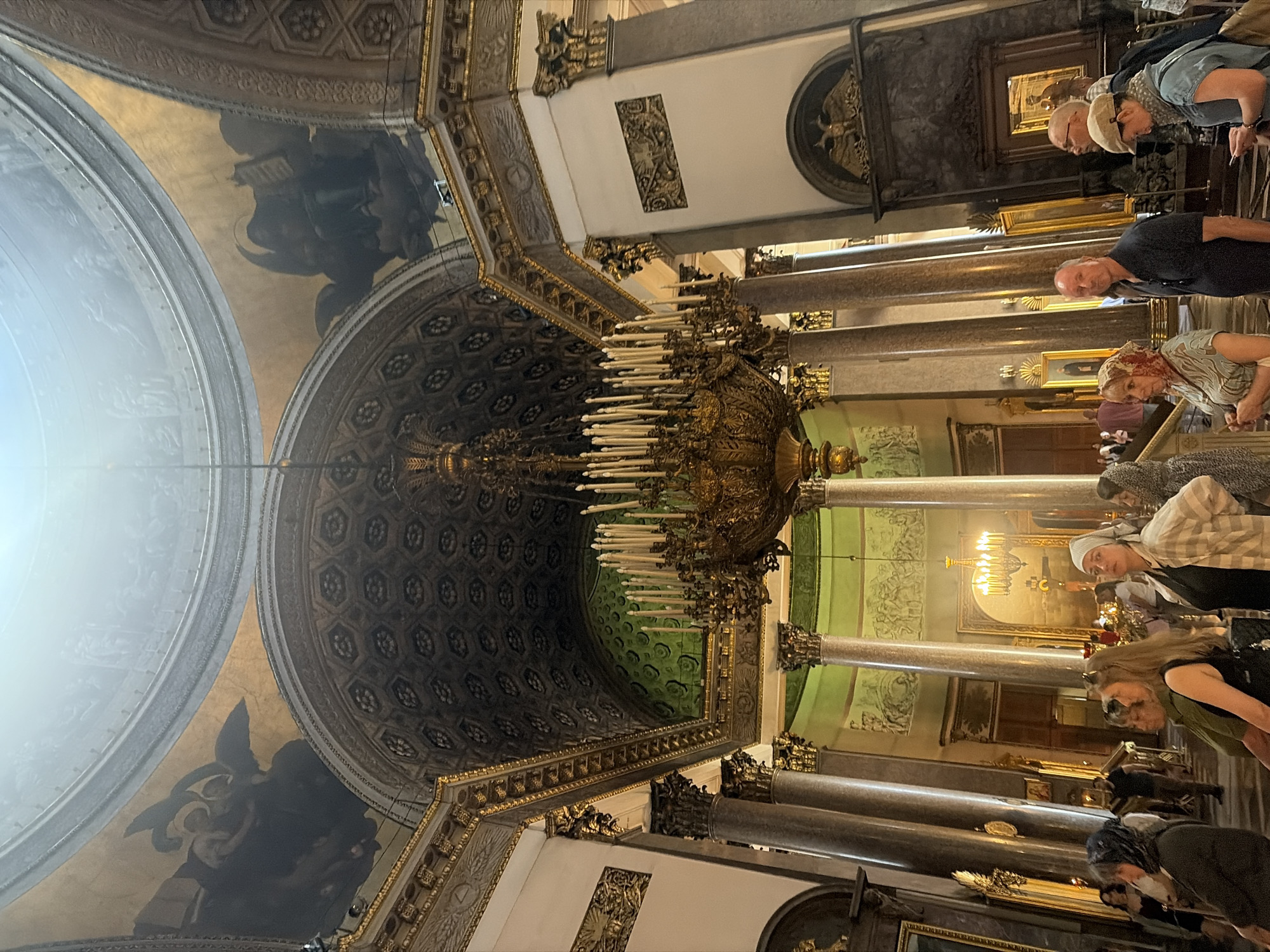 The image shows the interior of a large, ornate building, likely a cathedral or church, with a high, domed ceiling adorned with frescoes. A massive, multi-tiered chandelier with numerous candles hangs from the center of the dome. The walls are decorated with elaborate carvings and paintings, and large marble columns support the structure. Several people are present, standing and observing the architecture. Some are looking up at the ceiling, while others are engaged in conversation. The overall impression is one of grandeur and historical significance.
