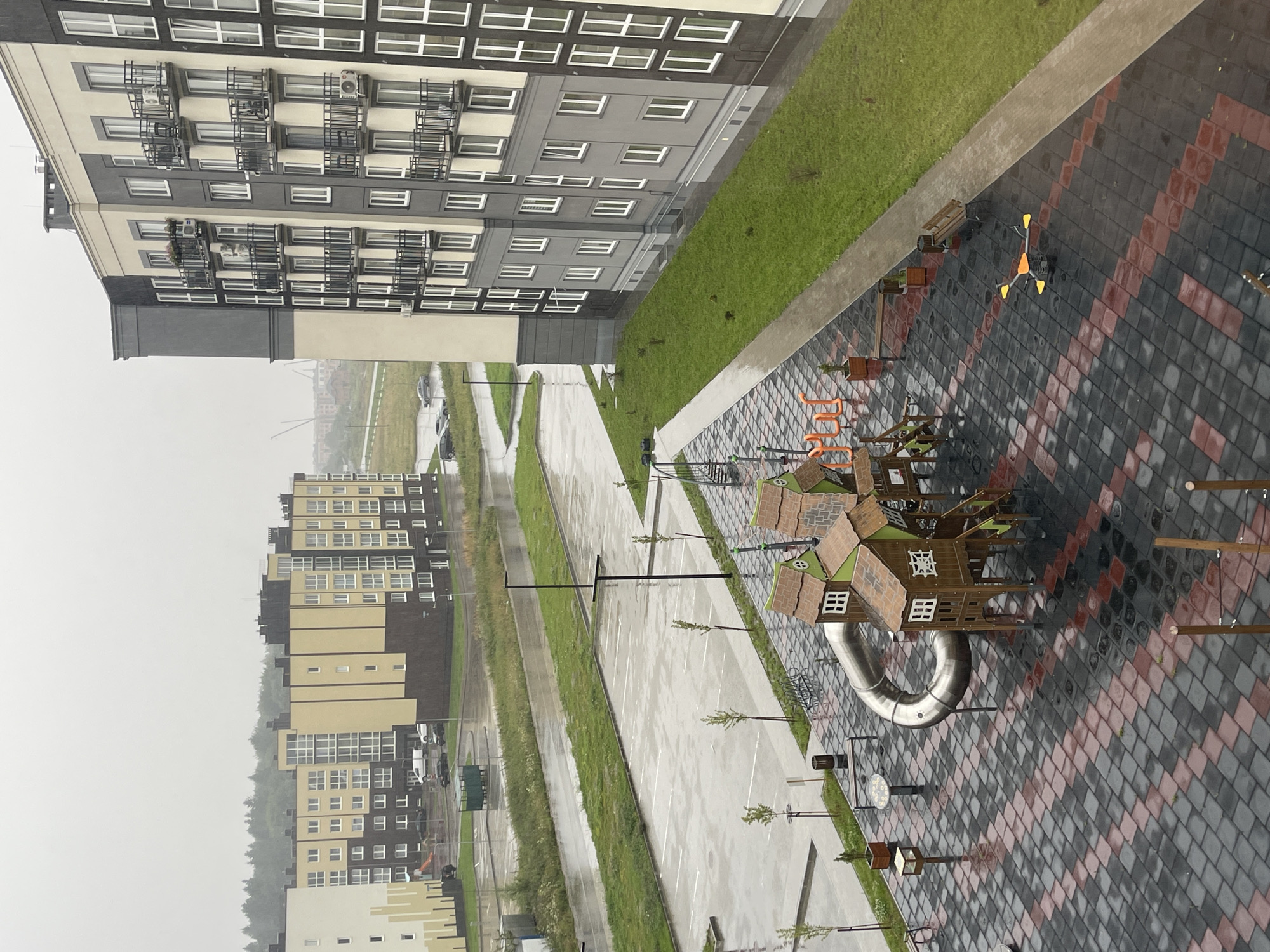 The image depicts a contemporary residential complex in Zelenogradsk, Russia, on a cloudy, rainy day. The foreground features a wet, patterned pavement of grey and red interlocking tiles, characteristic of modern urban landscaping. A children's playground is prominently visible, with a large, curved stainless steel slide, wooden playhouses with pitched roofs and windows, and colorful climbing structures. A yellow and orange seesaw-like spring rider is also present. Benches are strategically placed around the playground area.
In the mid-ground, a paved parking lot, also wet from the rain, is lined with white parking bays. A green refuse container is situated near the parking lot. Beyond the parking lot, a strip of green grass and sparse young trees separates it from a road. Several cars are parked on the road, and a white van is visible.
The background is dominated by a row of multi-story apartment buildings. These buildings exhibit modern architectural styles with a combination of light beige, grey, and dark facades, featuring numerous windows and balconies with black railings. Some buildings have visible air conditioning units and flower boxes on the balconies. In the far distance, construction cranes are visible, suggesting ongoing development in the area. The overall atmosphere is damp and overcast, with visible rain streaks on the pavement and buildings.