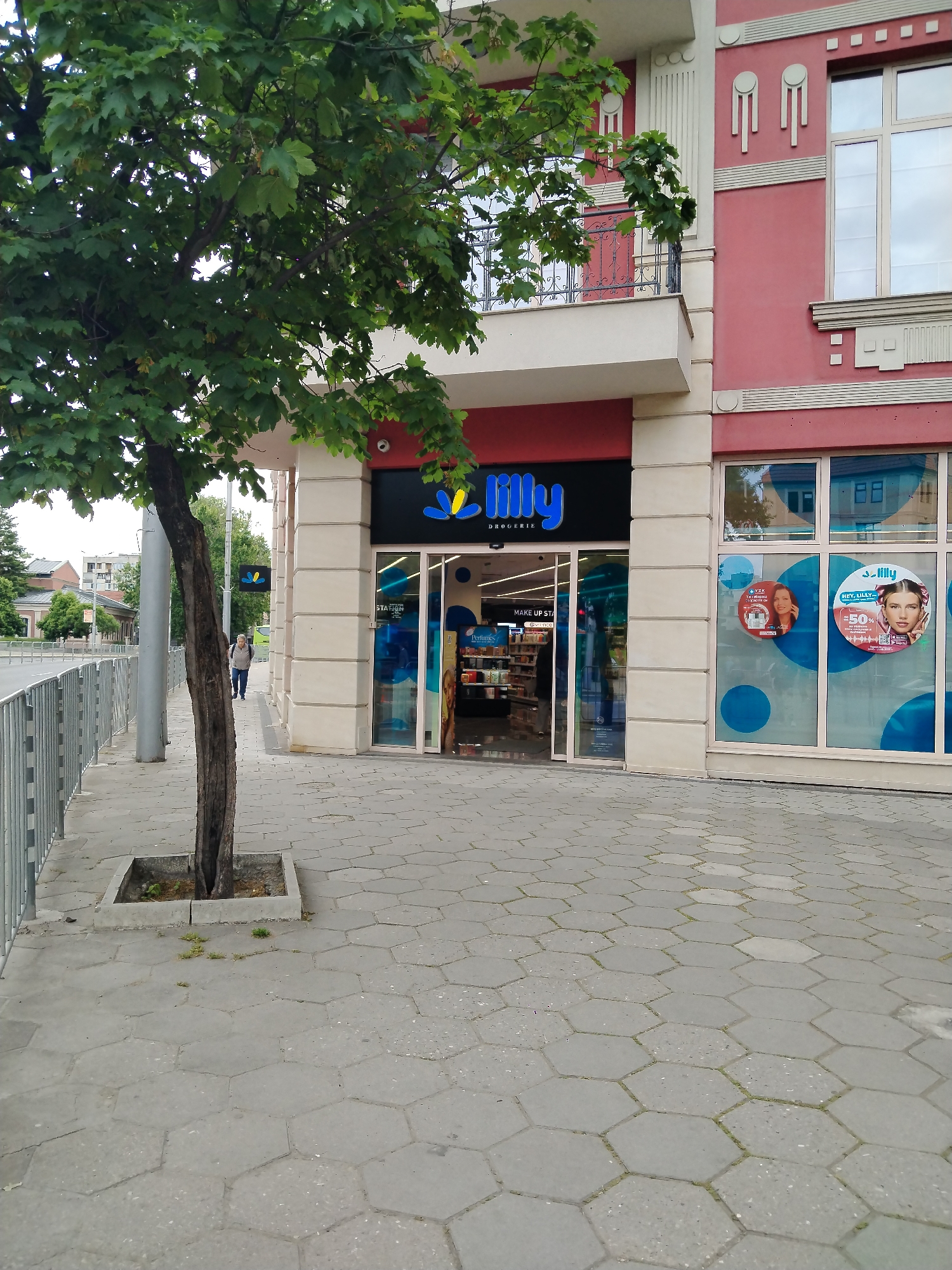 The image shows the exterior of a Lilly Drogerie store. The store is located in a city; based on the architectural style it is likely located in Plovdiv, Bulgaria. The building is a combination of cream and light red brick. The store has a black sign with the word "Lilly" in blue and yellow lettering. The entrance is flanked by cream-colored pillars and glass windows displaying advertisements. A tree with green leaves is on the sidewalk in front of the store, planted in a square concrete container. The sidewalk is paved with grey hexagon-shaped stones. A pedestrian is walking down the sidewalk past the store. A metal fence is visible along the side of the sidewalk.