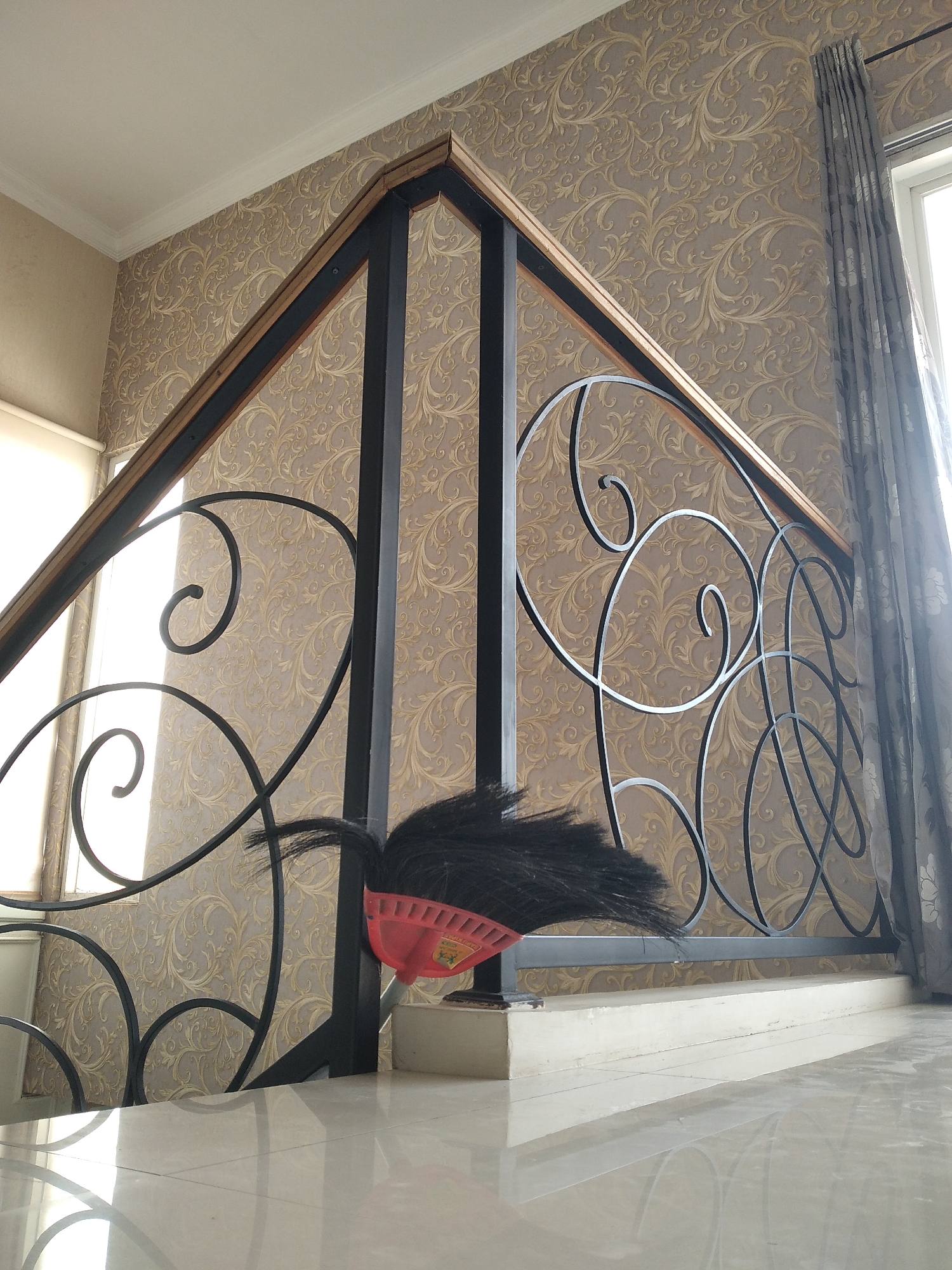 The image captures a close-up view of a staircase railing with a decorative pattern, set against a wall adorned with ornate wallpaper. The railing is made of black metal with a curved, scrolled design and has a wooden trim along the top. A red broom with black bristles is leaning against the railing, partially obscuring the lower portion of the design. To the right of the railing, a gray curtain hangs near a window, and the floor in the foreground is tiled and highly reflective. The lighting suggests it is daytime, with natural light coming from the window. The scene appears to be an interior, possibly a home or a building in Kaliwates, Indonesia, where such architectural and decorative elements are common.