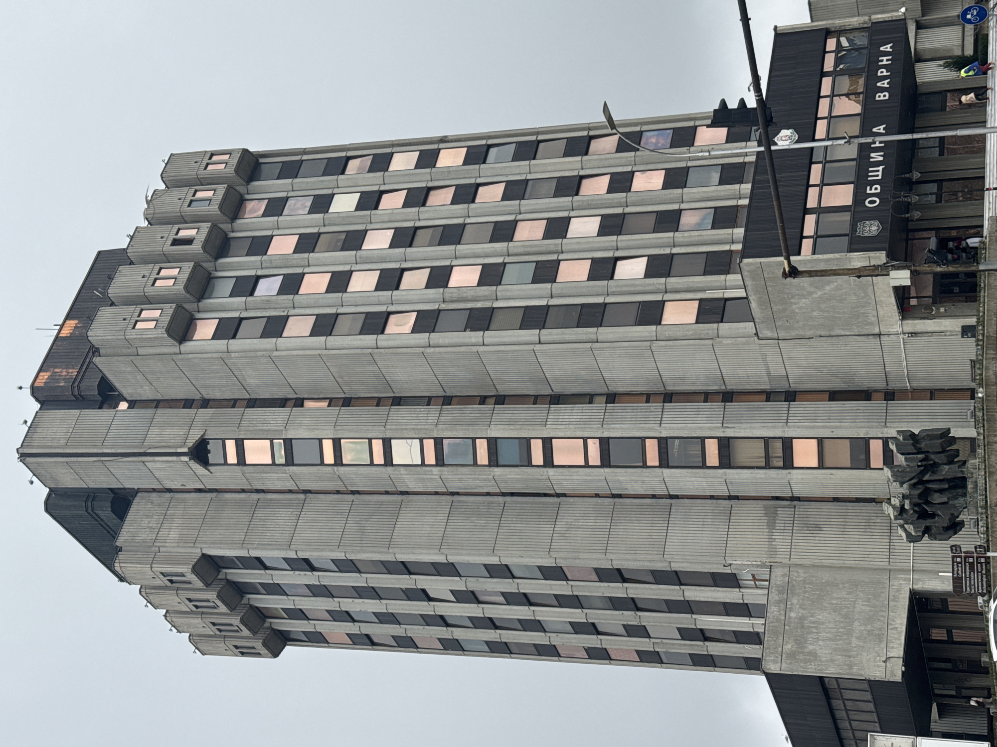 The image shows a tall, brutalist building in Varna, Bulgaria. The building is constructed of concrete and has a repetitive pattern of windows. A sign on the side of the building reads "Община Варна" (Municipality of Varna) and "Varna." The sky is overcast. There are people visible near the entrance. A sculpture and street signs are in the foreground.