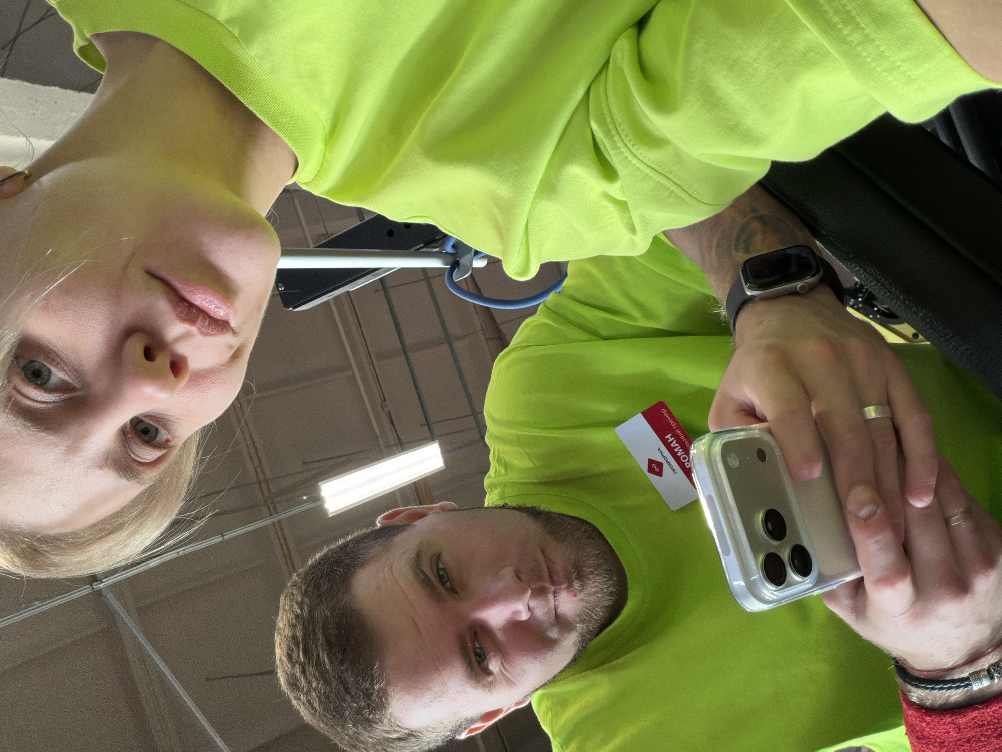 A low-angle selfie captures two individuals wearing bright lime green shirts, against a backdrop of an indoor ceiling with fluorescent lighting. The person on the left has fair skin and blond hair. To the right, the second person has darker hair and a beard, and is holding a smartphone with a clear case. A white card is visible between the smartphone and their shirt. On the wrist of the smartphone-holding person, there is a silver smartwatch and a braided bracelet.
Although the location can not be confirmed, the card held by the person states "ПРОФСОЮЗНЫЙ ТРЕКЕР" ("Trade Union Tracker") and contains text in Cyrillic. Given this information, it is plausible that the image was taken in Russia. If it were taken in Chaikovsky, Russia, it would likely be in an indoor location.