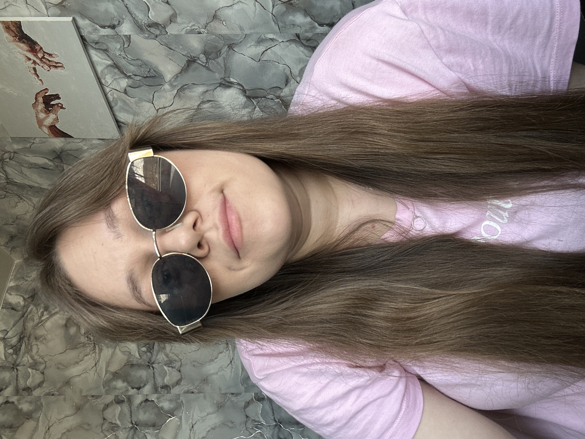 A young woman with long brown hair is wearing gold-rimmed sunglasses and a pink t-shirt. She appears to be lying down. In the background is a marbled gray wall and a framed print depicting hands reaching towards each other. The location is stated as Ольховка, Russia, but there is no verifiable information within the image to support this.