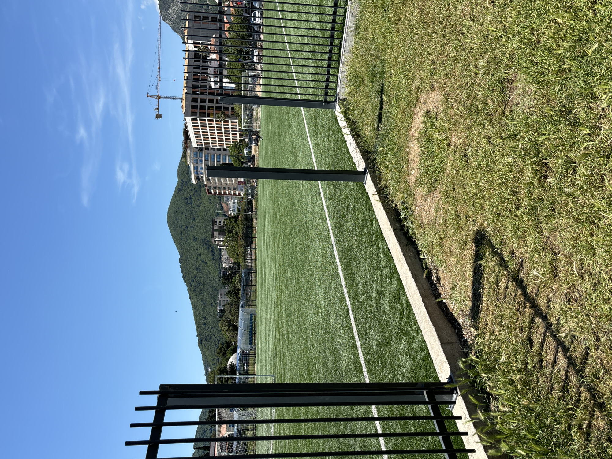 The image shows a fenced soccer field with artificial turf in Budva, Montenegro. A mountain backdrop is present, and modern buildings, including one under construction with a crane, are visible. Grass grows on the opposite side of the fence from the field. The sky is clear and blue.