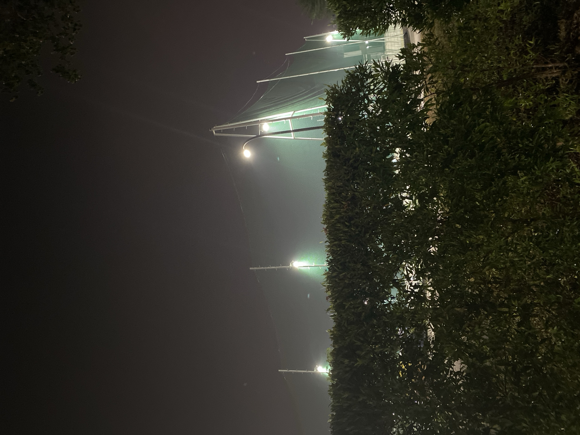 The image depicts a night scene at a golf driving range in Tangerang, Indonesia. A dense green hedge runs across the foreground, obscuring the lower portion of the scene. Behind the hedge, tall poles support a large green netting, characteristic of a golf driving range. Several bright floodlights are visible, illuminating the netting and the surrounding misty atmosphere. The sky is dark and overcast. The overall impression is one of a sports facility operating at night.