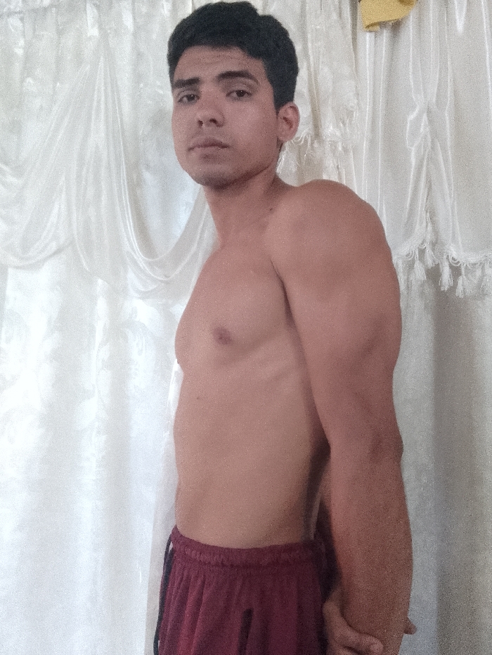 A young adult male, appearing in his late teens or early twenties, stands shirtless indoors in Juan Griego, Venezuela. He possesses short, dark hair and a muscular build, visible in his chest, shoulders, and arms. His gaze is directed forward towards the camera with a neutral expression. He is wearing maroon athletic shorts with a dark vertical stripe along the side. His left arm is positioned slightly forward, while his right forearm and hand are visible towards the bottom right, with his hands clasped or interlocked. The background consists of light-colored, possibly white or cream, fabric curtains with visible drapes, folds, and subtle patterns, consistent with an interior residential setting. A small yellow object is partially visible at the top right of the frame, against the curtain.