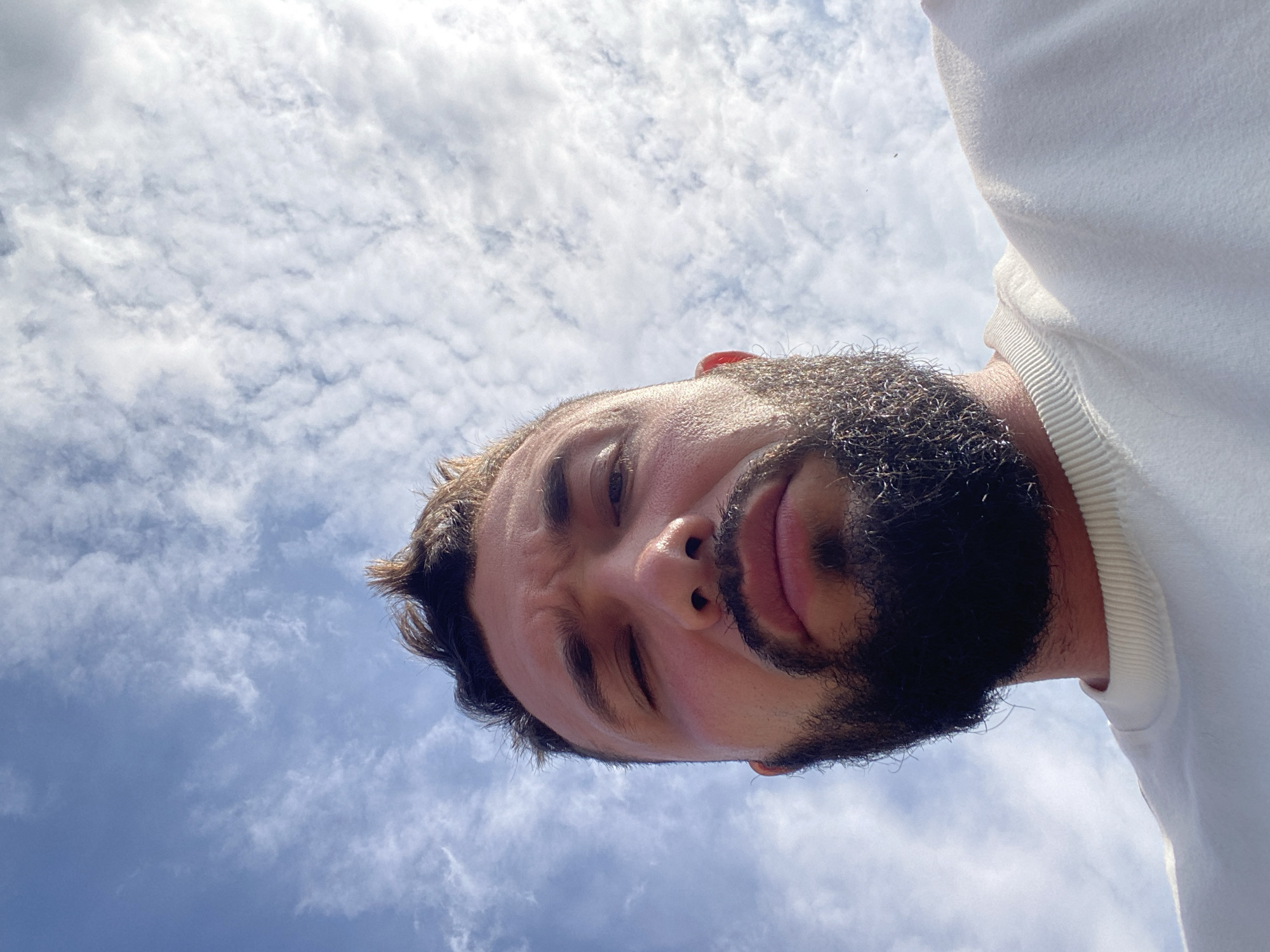 A low-angle, close-up, outdoor selfie shows a man looking up at the camera. The man has dark, short, spiky hair and a full, dark beard and mustache. His eyes are visible, with his left eye appearing to be partially closed. He is wearing a white crewneck sweatshirt. The background is a bright blue sky with scattered white clouds. The lighting suggests it is daytime, possibly sunny. There are no identifiable landmarks or features that would indicate the location in Chalda, Russia.