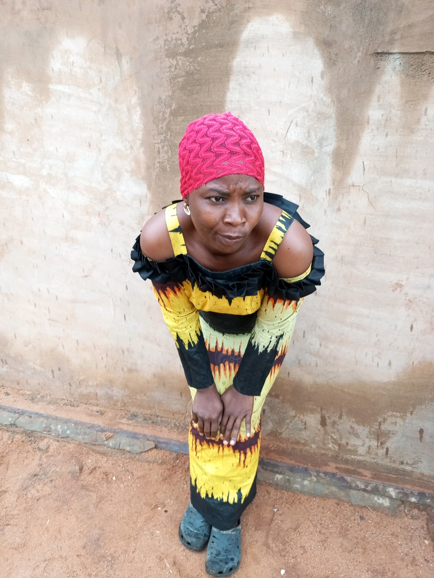 A woman with dark skin, wearing a bright pink headscarf and a yellow, black, and orange patterned dress, is crouched down in front of a light brown, textured wall. Her hands are resting on her knees, and she is looking slightly upwards and to her left with a serious expression. Her dress has off-the-shoulder sleeves with black ruffles. She is wearing dark blue clogs. The ground in front of her is dirt and sand. The scene appears to be outdoors, possibly in a rural or less developed area of Tempure, Nigeria.