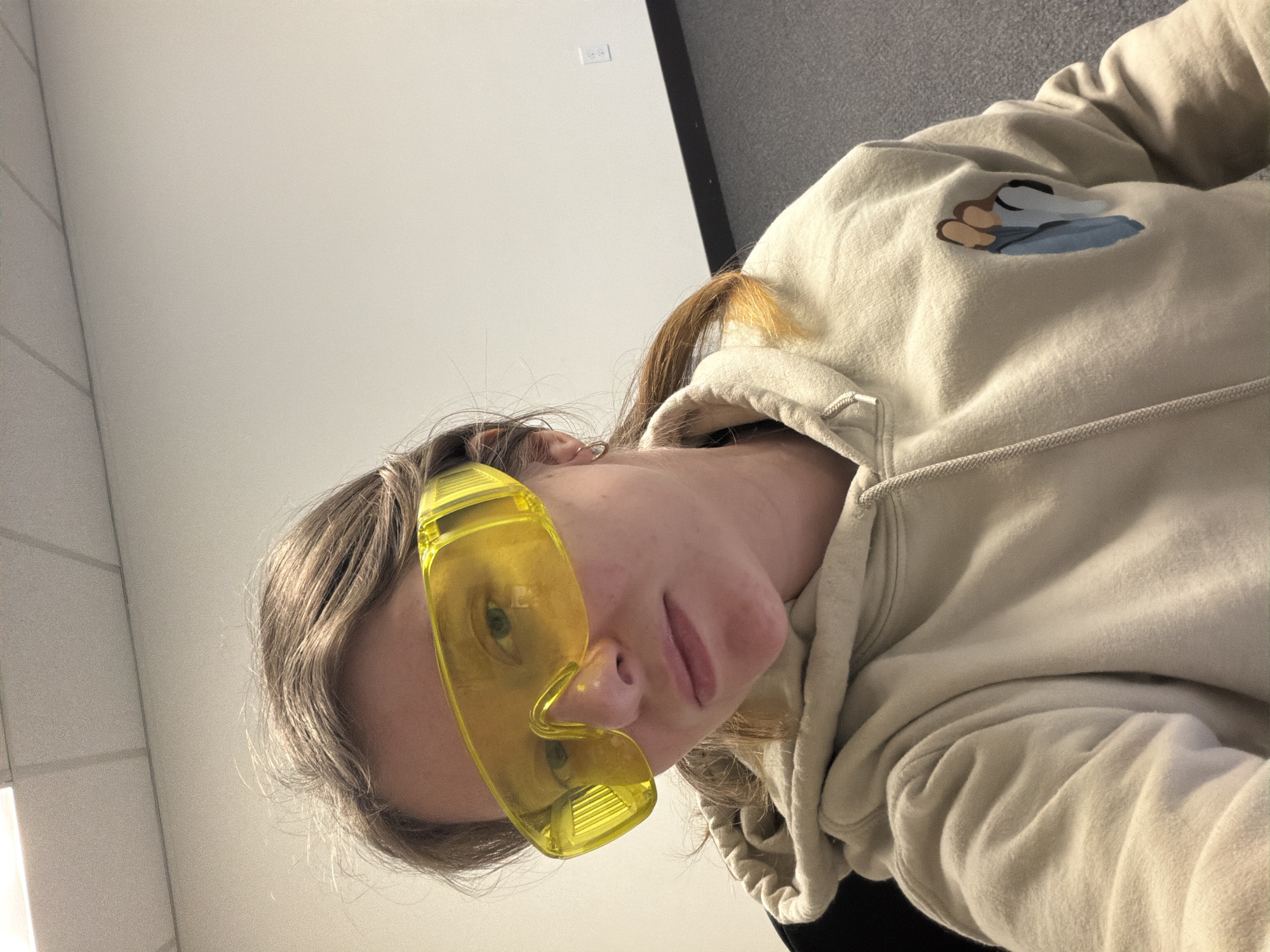 This is a vertically oriented, close-up, selfie-style photograph of a young person. The person is wearing a light tan hooded sweatshirt with a graphic of two figures in blue and brown on the left chest. They are also wearing bright yellow safety glasses that cover a significant portion of their face, obscuring their eyes and nose. Their mouth and chin are visible, and they have light brown hair pulled back into a ponytail that is visible on top of their head. The background is out of focus, but appears to be an indoor setting with a white wall on the left and a darker, textured surface on the right. A light switch is visible on the white wall. The lighting suggests it is daytime. The person appears to be relaxed and perhaps playful, given the unusual choice of safety glasses for a selfie. There is no visible text in the image. The location context is Beaverton, United States of America.