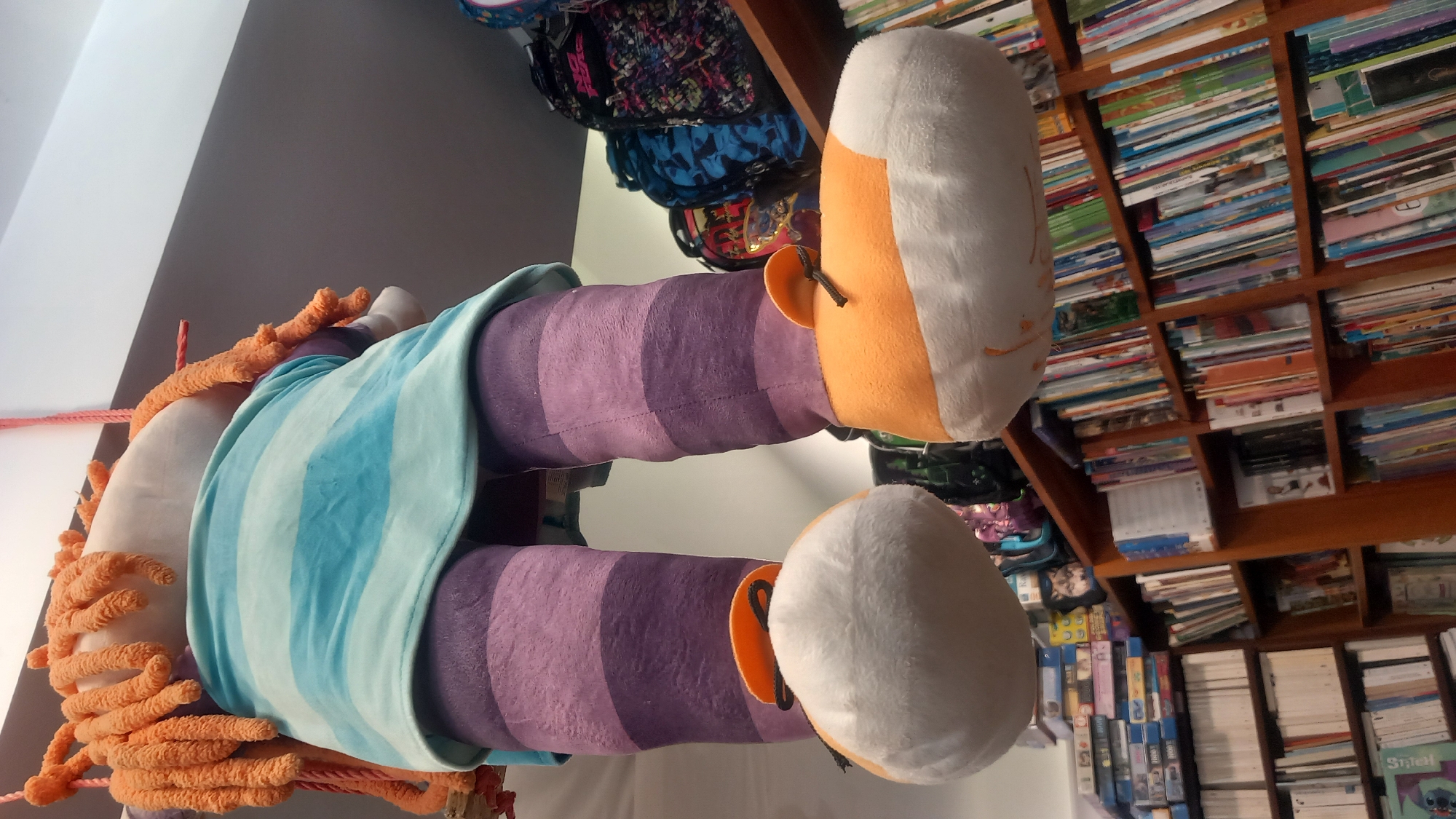 The image shows a large, plush doll hanging from a string. The doll has bright orange yarn hair, a light blue and white striped dress, and purple and purple-striped legs. Its feet are large, white, and orange plush shoes. The doll is hanging in front of bookshelves filled with books. In the background, on a shelf above the books, are several backpacks. The overall scene suggests a toy store or a similar retail environment, possibly within the city of Kos, Greece, though the location cannot be definitively confirmed from the image alone. The lighting suggests an indoor setting, with artificial light sources.