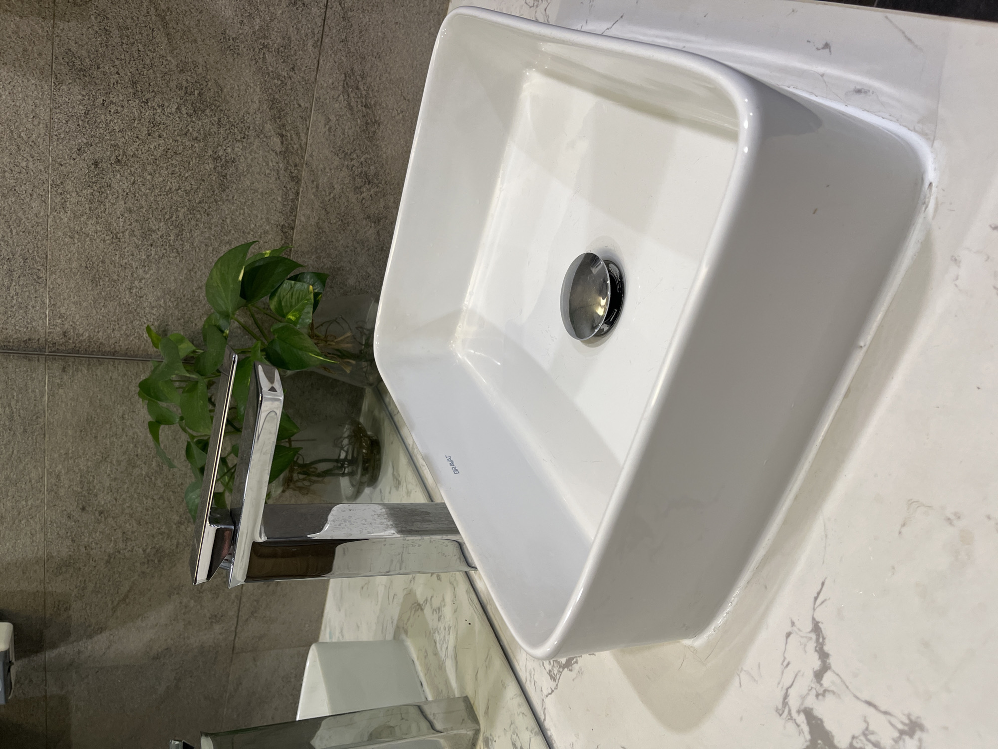 The image shows a bathroom sink. The sink is white, square-shaped, and appears to be made of ceramic. It is installed on a countertop that has a white and grey marble pattern. A chrome faucet is visible to the left of the sink. A small green plant in a clear vase is positioned behind the faucet. The background features a grey, textured wall. The specific location in Đà Nẵng, Vietnam cannot be determined based on the visual information provided.
