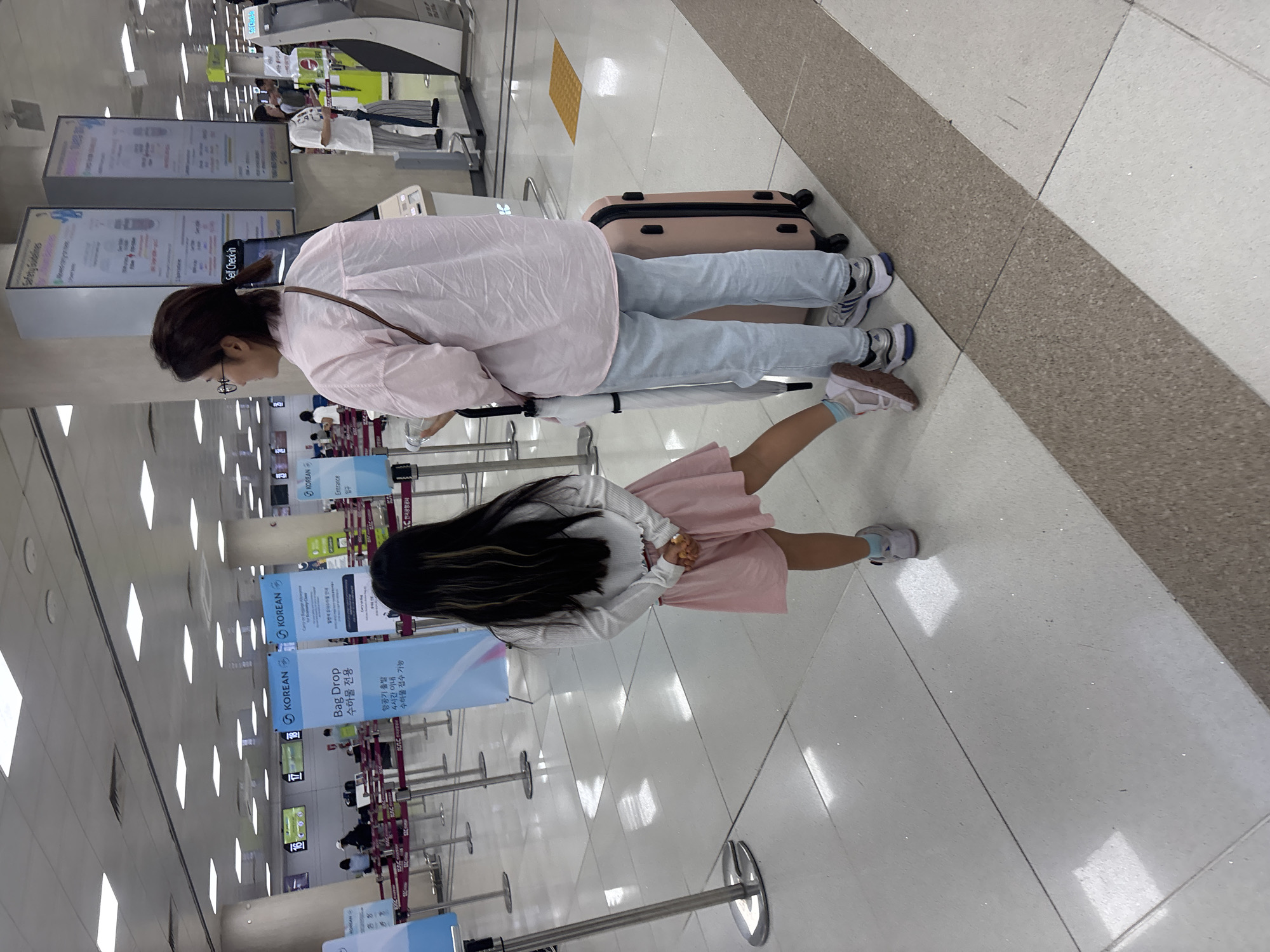 An exterior wide shot captures a mother and daughter standing in an airport terminal. The mother, a woman with dark hair pulled back, wears glasses and a light pink oversized button-down shirt with light blue jeans. She holds a water bottle in her left hand and stands next to a light pink suitcase and a white umbrella. The daughter, with long dark hair, wears a white long-sleeved top and a pink skirt, her hands clasped behind her back. They are positioned in a tiled airport hallway with various signs in Korean and English, indicating a "Bag Drop" area. The environment is well-lit with ceiling lights and displays visible in the background. The airport terminal is the departure hall of Jeju International Airport, located in Jeju City, South Korea.
