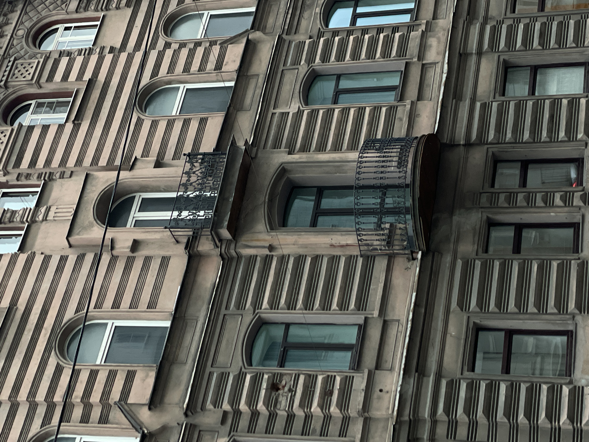The image shows a section of an architectural facade in Saint Petersburg, Russia. The building is characterized by vertical striping and alternating window shapes; some are arched, and others are rectangular. One window has a small wrought-iron balcony. The building's construction and design are indicative of a historical architectural style.