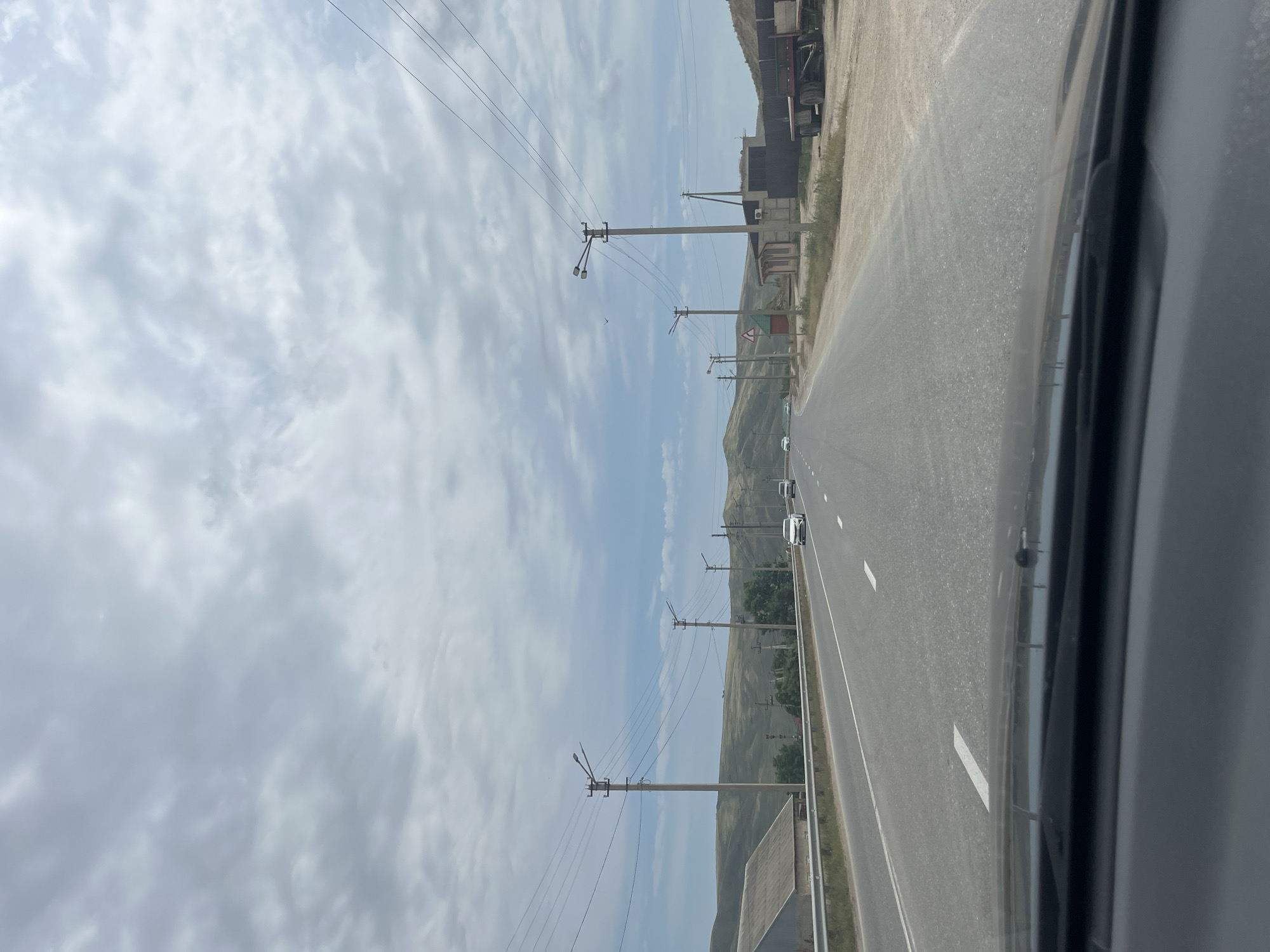 The image shows a highway on a cloudy day, with rolling hills in the background. The road is asphalt and appears to be in good condition, with white lane markings. There are several cars on the road, and utility poles with power lines line the sides of the highway. A building with a gray roof is visible on the left side of the road, and a red truck is parked on the right side. The sky is filled with gray and white clouds, indicating overcast weather. The overall scene suggests a rural or suburban setting outside of Karabudakhkent, Russia.