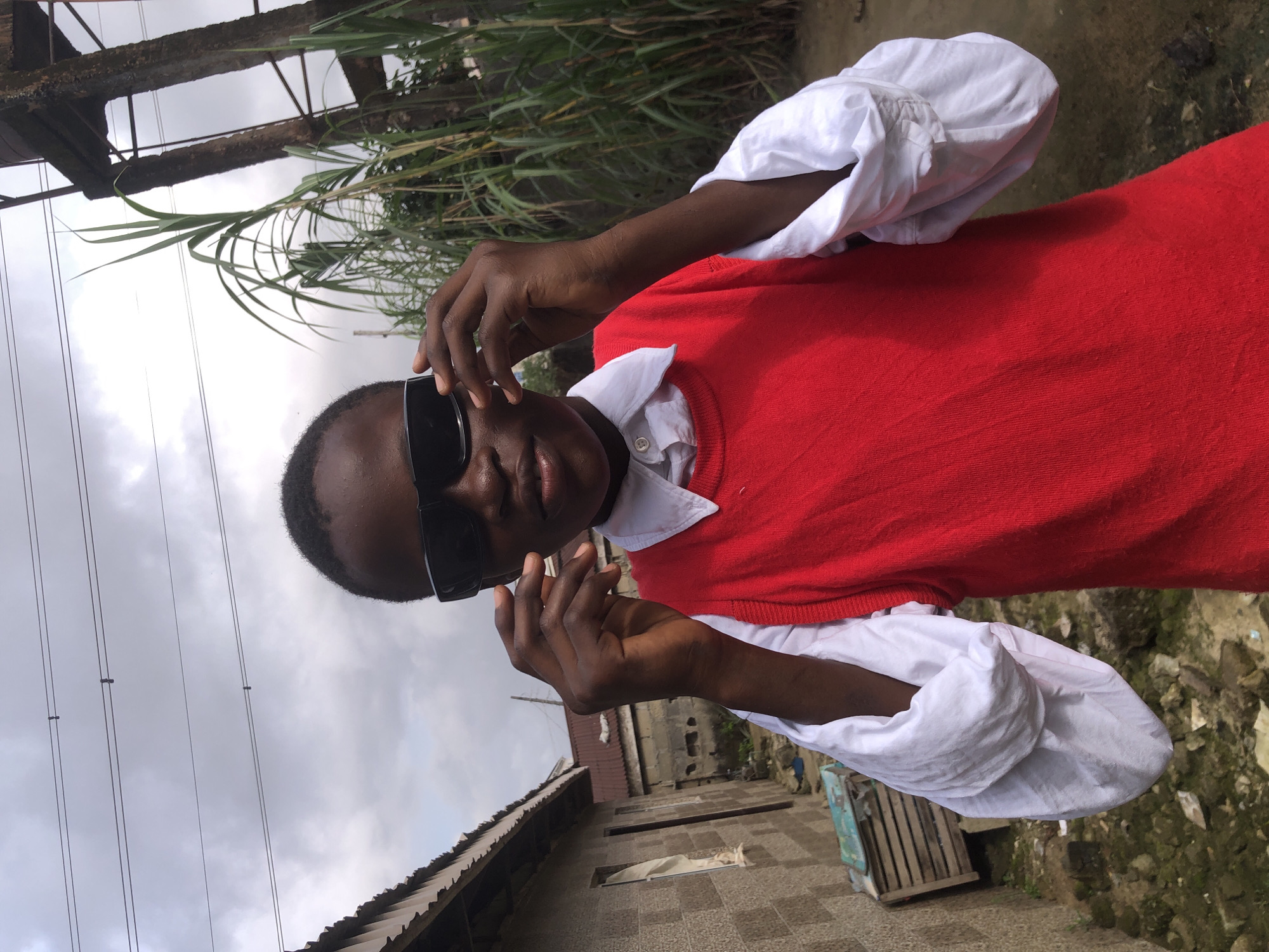 In Ado, Nigeria, a young person wearing sunglasses and a red vest over a white collared shirt poses outdoors. Their hands are adjusting the sunglasses. In the background, there is a grey brick building with a corrugated metal roof, tall green plants resembling sugarcane, a water tower or similar structure, and power lines against a cloudy sky. The ground is uneven and appears to be dirt with some debris.