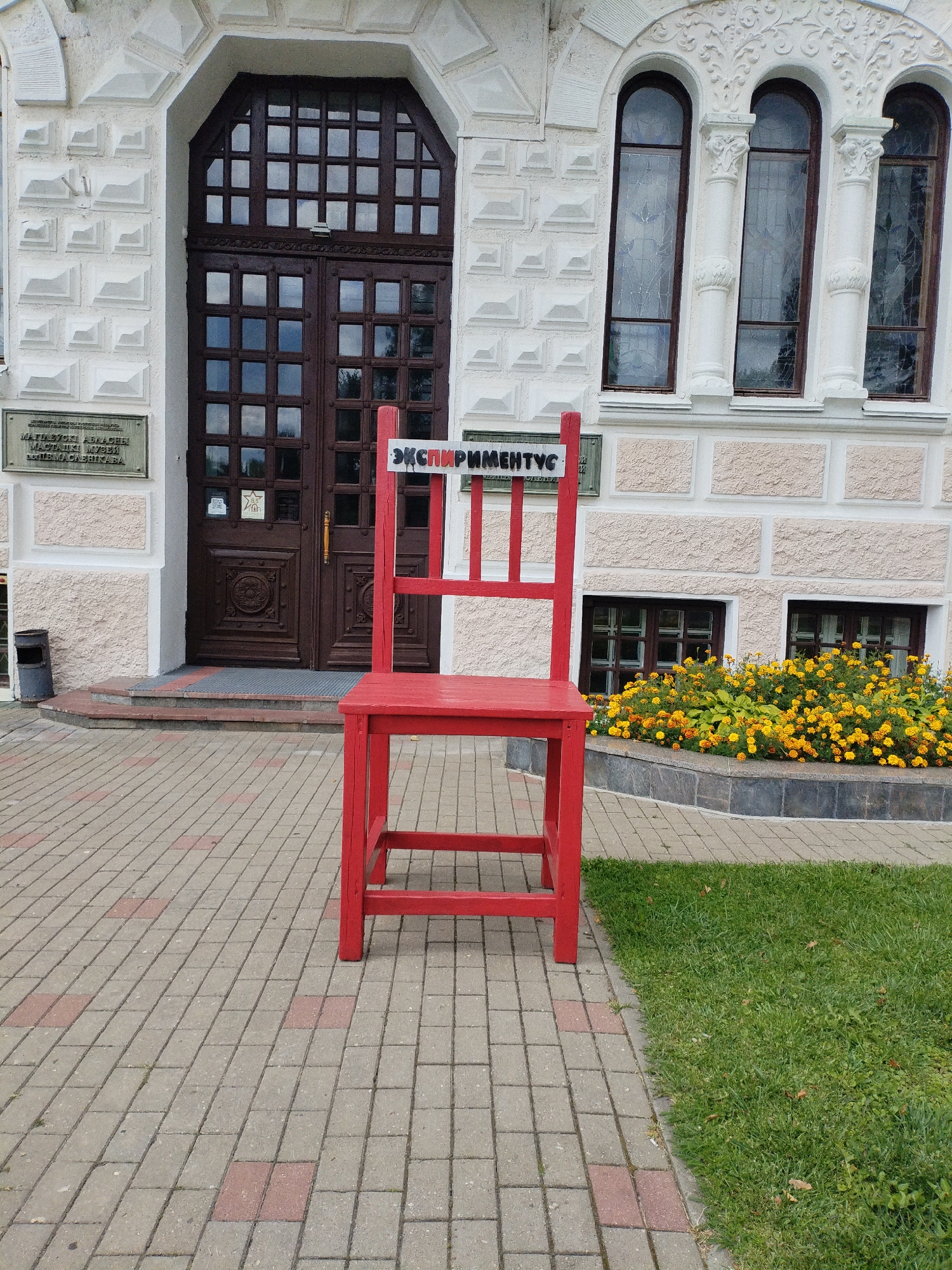A large, bright red chair is positioned outdoors on a brick-paved walkway in front of a building with a dark brown wooden double door. The building features ornate white architectural details, including a decorative archway above the door and arched windows with stained glass. A sign in Russian is visible on the wall to the left of the door, and another sign with Cyrillic lettering is affixed to the red chair. A flower bed with yellow and orange flowers is located to the right of the chair, in front of a window. The ground is paved with rectangular bricks, interspersed with some red bricks. Lush green grass borders the walkway and flower bed.