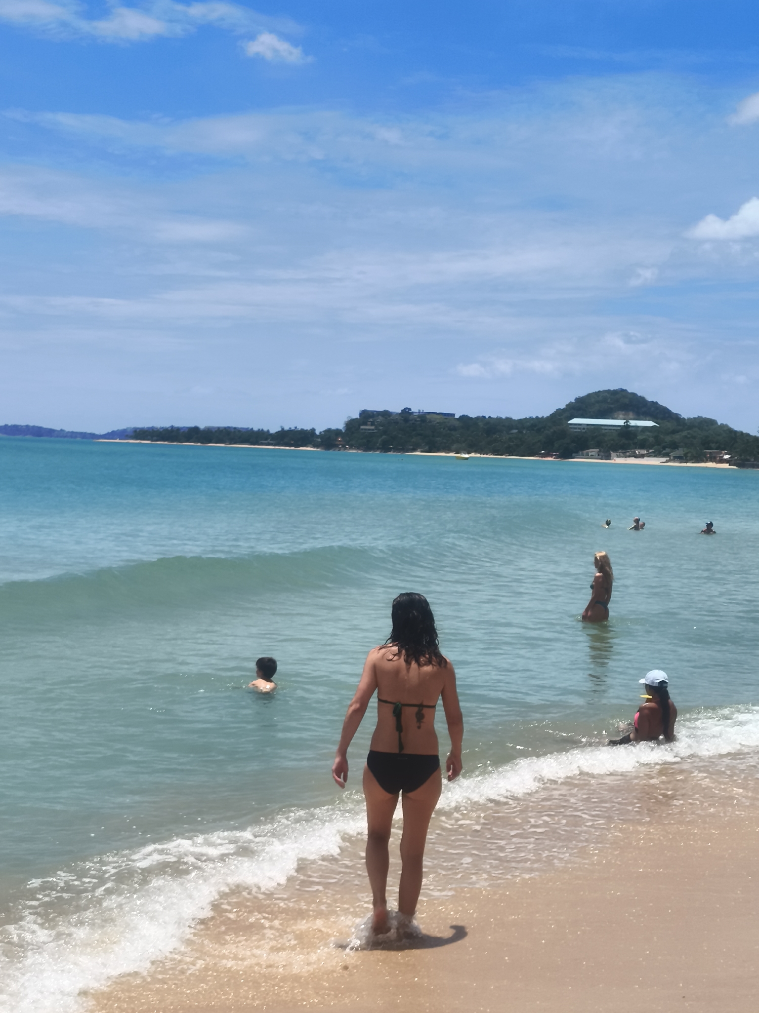 An eye-level, wide-angle shot captures a beach scene in Ko Samui District, Thailand. The turquoise ocean meets a sandy shore under a bright blue sky dotted with white clouds. In the foreground, a woman with dark hair in a black bikini stands facing away from the camera, her feet touching the foamy edge of a wave. Several other people are in the water. To the left, a young child with dark hair is swimming. Further out, a blonde woman in a dark bikini stands in the water. To the right, a person wearing a white cap and a dark swimsuit sits near the shore. In the background, a lush green island with a large building on its summit forms the horizon.