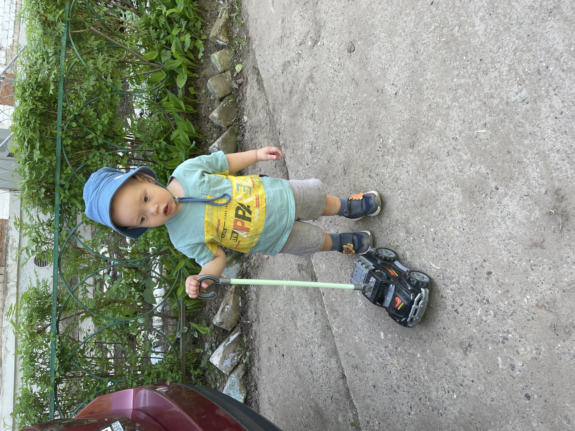 A young child is captured in an outdoor setting, pulling a black toy truck. The scene is bright, suggesting daytime. The child, likely a toddler, is wearing a blue bucket hat, a light green t-shirt with yellow accents and text, grey shorts, and navy blue shoes with orange details. Their expression is neutral, looking slightly towards the camera.
The child holds a grey handle attached to a light green pole, which connects to the black toy truck. The truck has a rugged design with orange-red accents and grey bumper-like features. It has four wheels and appears to be a pull-along toy.
In the background, to the child's left, is a dense line of green bushes and foliage, partially obscured by a green metal fence or railing. The ground is a light grey concrete or paved surface. Part of a dark red or maroon car is visible in the lower left corner of the image. A blurry license plate on the car shows "B 133 BA 59 RU", indicating the Perm Krai region of Russia.
Visible text on the child's t-shirt includes phrases like "ORIGINAL JEANS APPAREL EST. 1990," "CASUAL DIVISION," and "PREMIUM LEGENDS," along with some large, partially visible letters.