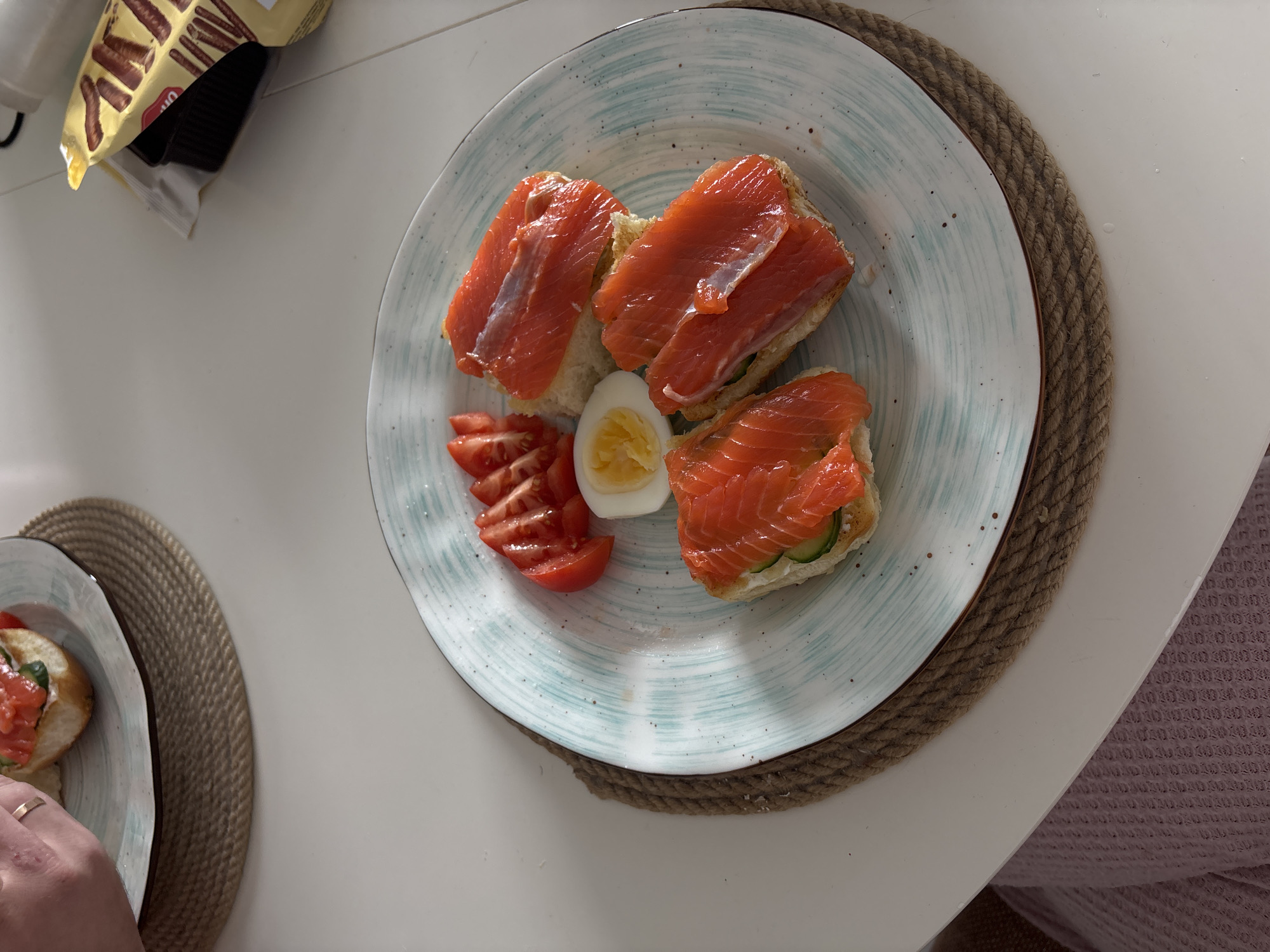 A top-down shot presents a light blue and white plate holding a composed meal. The plate contains three open-faced sandwiches topped with salmon, arranged in a triangular configuration. Sliced tomatoes and half of a hard-boiled egg are positioned on the left side of the plate. The plate rests on a round, woven brown placemat. To the upper left of the plate, a partially visible package of chocolate-covered biscuit sticks sits on a white table. Another plate with a meal is partially visible on the lower left. A hand, wearing a ring, is also partially visible in the lower left corner. The table appears to be located indoors, with a purple waffle weave fabric visible on the right. The location cannot be determined as Krasnoyarsk, Russia based on the visual data.