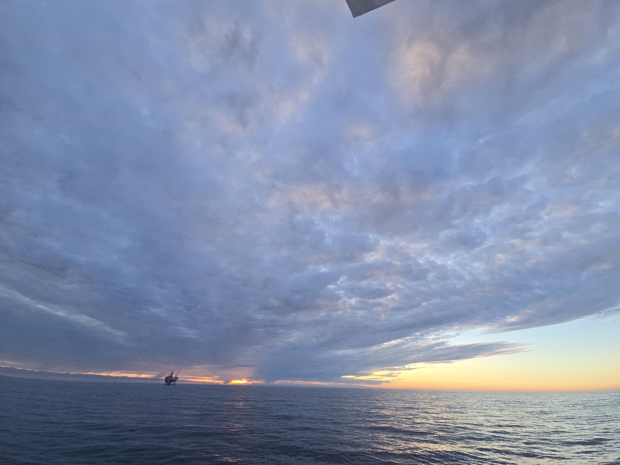The image shows a wide expanse of ocean under a cloudy sky. The clouds are predominantly gray and blue, with some patches of pink and white. Near the horizon, the sky transitions to a yellow and orange hue, suggesting a sunrise or sunset. On the left side of the image, a silhouette of an offshore oil platform or similar structure is visible on the horizon. The ocean is dark blue and displays gentle ripples on its surface. The location is an unknown ocean location in the city of Unknown, Unknown.