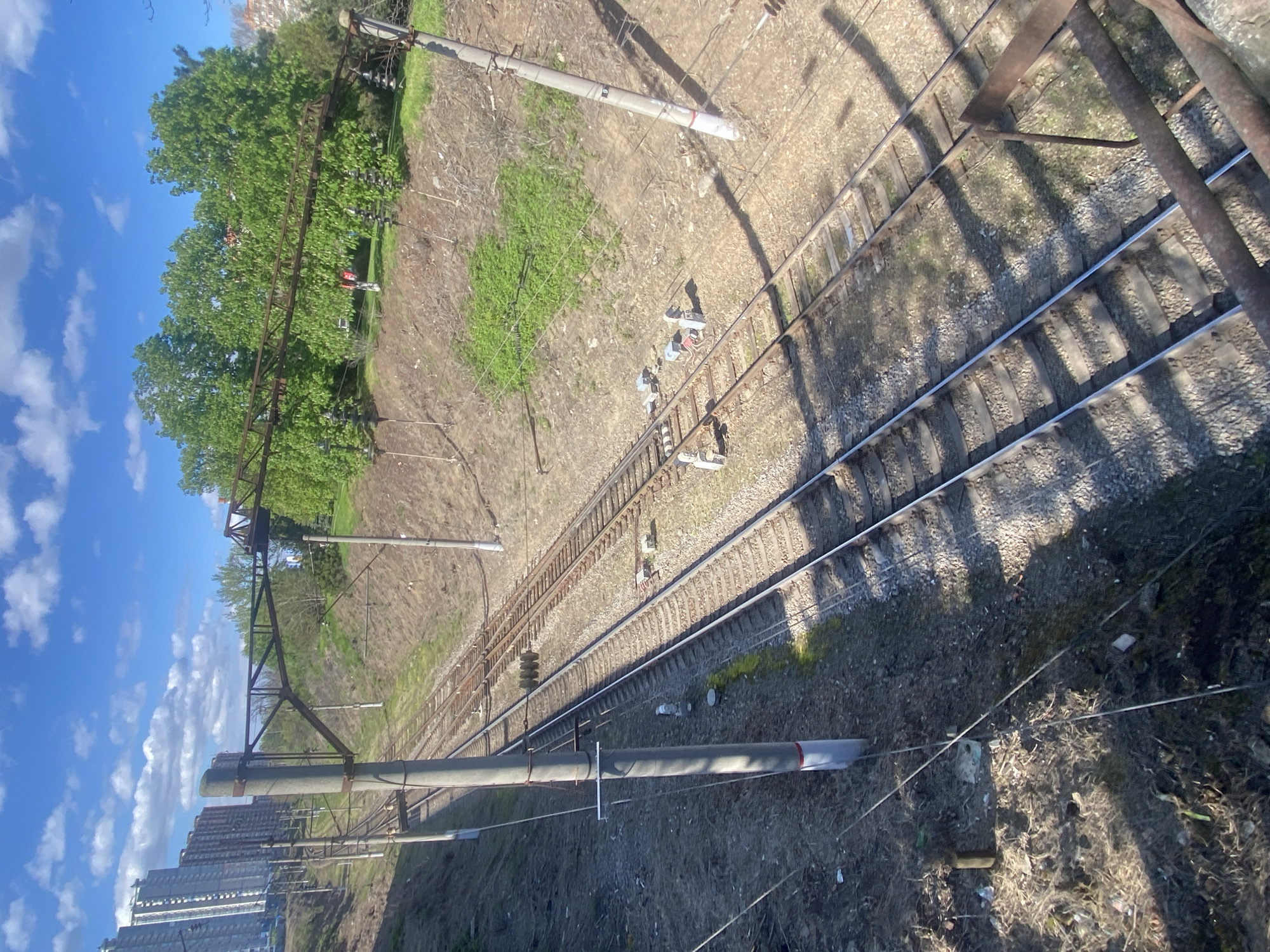 The image depicts a panoramic view of multiple railway tracks set in an urban or suburban environment under a clear, sunny sky. The perspective is from above, looking down diagonally along the length of the tracks.
In the foreground and extending into the mid-ground, several parallel railway tracks are visible, complete with sleepers and gravel ballast. Overhead power line structures, consisting of tall concrete poles and intricate metal frameworks supporting numerous wires and insulators, stretch across the scene, indicating an electrified railway system. Some small, white or light-colored signal boxes or equipment modules are positioned at intervals beside the tracks. The ground adjacent to the tracks is mostly dry earth and sparse, dry grass, with some patches of greener vegetation and scattered litter in places.
The background on the left side of the frame features a lush line of green trees, beyond which several multi-story buildings, possibly residential, are visible under a bright blue sky dotted with white clouds. On the right, the tracks continue into the distance, bordered by more undeveloped, grassy terrain. Long, distinct shadows cast by the overhead structures across the tracks and ground suggest either a mid-morning or late afternoon time of day.
There are no clearly discernible people or animals present in the scene. A very small, indistinct red object, possibly a distant figure or piece of equipment, is visible among the trees in one of the cropped sections. No visible text is present in the image. The overall impression is one of a functional railway infrastructure integrated into a semi-urban landscape.