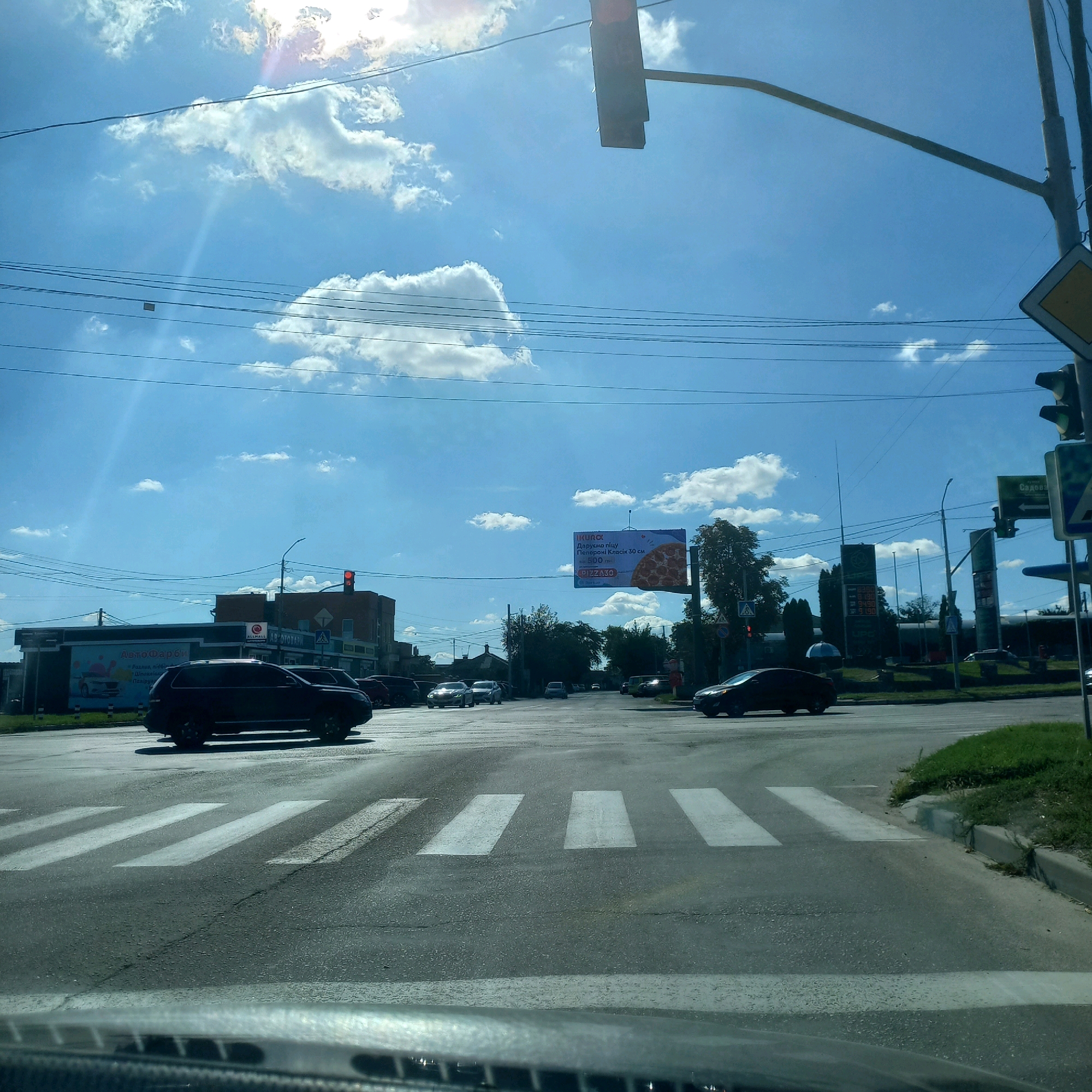 The image depicts a view from a vehicle, looking forward towards a street intersection in Kropyvnytskyi, Ukraine. The sky is bright blue with scattered white clouds, and the sun is visible, casting lens flare. In the foreground, a crosswalk is marked on asphalt. Several cars are visible on the road, including a dark SUV crossing the crosswalk from the left. Buildings line the street, with signs including "ALLMALL" and advertisements for pizza and auto paints. Traffic lights are present, with one appearing red. The environment suggests a typical urban street scene on a sunny day.