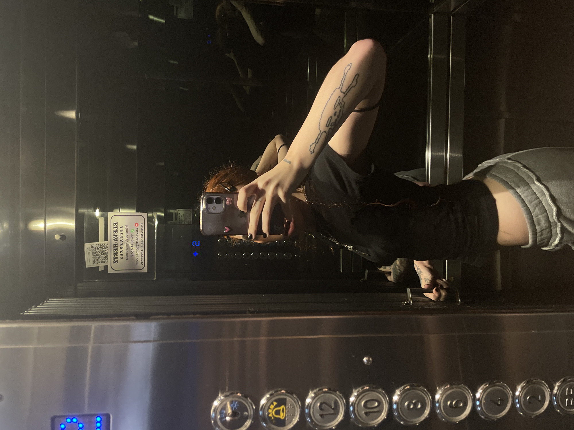 A person with reddish hair stands inside an elevator in Buenos Aires, Argentina, taking a selfie in the mirror. The individual has tattoos on their left arm. They are wearing a black t-shirt and gray shorts. The elevator has buttons for various floors, including the ground floor. There is a sign with a QR code and other information in Spanish on the wall of the elevator. The lighting is dim.