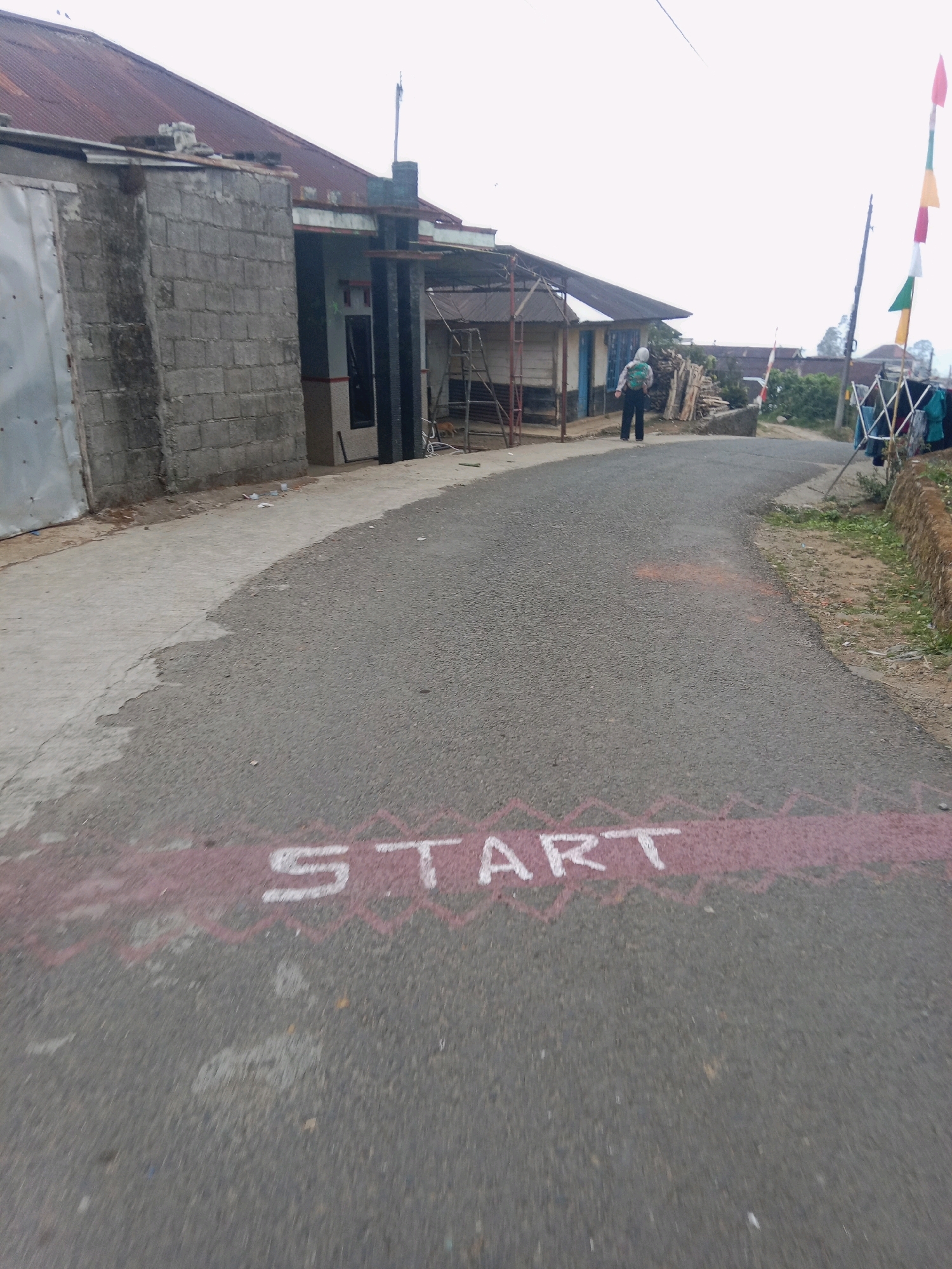 In Batur, Indonesia, on a rural road, the word "START" is painted in white lettering on the asphalt, bordered by a zig-zagged pink line. A person wearing a light-colored hooded jacket and dark pants, carrying a backpack, stands on the road facing away from the camera. The road curves to the right, leading towards a cluster of buildings and vegetation. To the left of the road, a brick building with a corrugated metal roof is visible, along with a concrete path. To the right, a clothesline with laundry, flags on poles, and a low stone wall line the roadside. The sky is overcast.