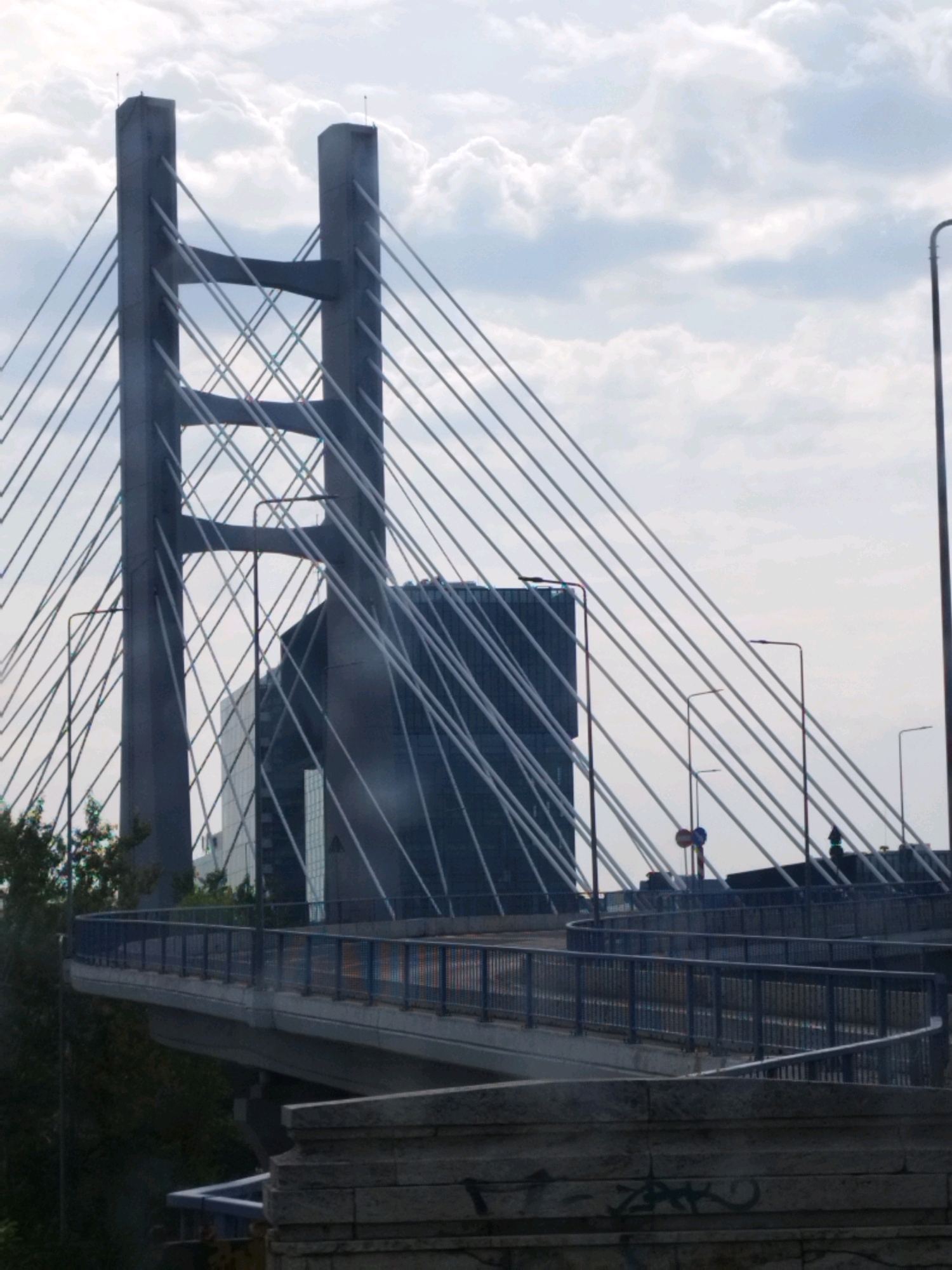 The image depicts the Basarab Bridge in Bucharest, Romania, a modern cable-stayed bridge. In the foreground, a section of the bridge's roadway with a blue railing is visible, curving to the right. Behind it, the bridge's large, grey concrete pylons rise, supporting numerous thin cables that fan out across the sky. A modern, dark glass-clad building with a rectangular shape stands behind the pylons, partially obscured by them. The sky is overcast with white and grey clouds. Several streetlights are visible along the bridge. In the distance, foliage is present on the left side of the image. The overall scene is one of urban infrastructure against a cloudy sky.