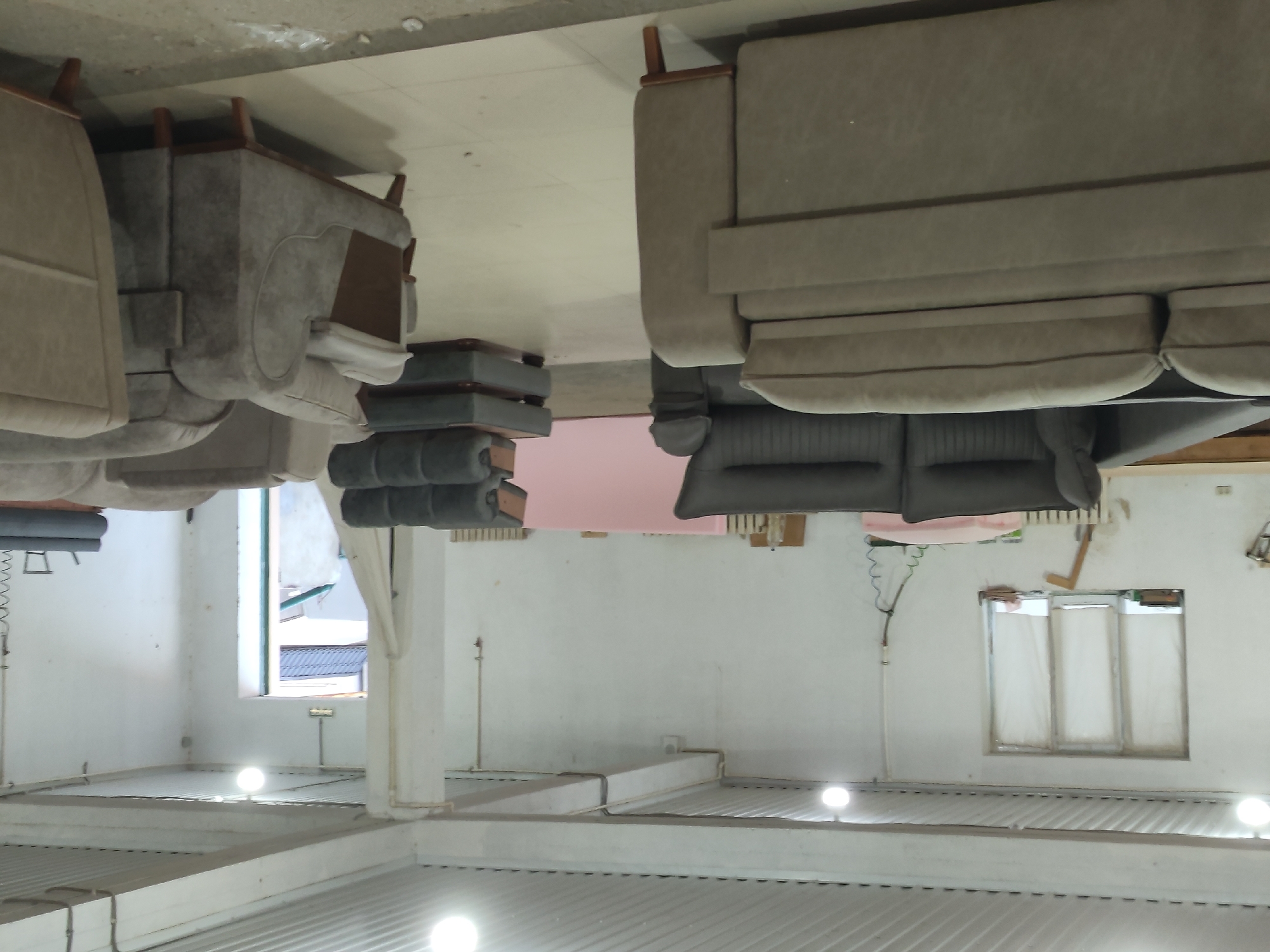 The image depicts the interior of a furniture workshop or showroom in Leninkent, Russia. The space is large and open, with a high, corrugated metal ceiling and white walls. Several pieces of upholstered furniture are visible, primarily sofas and armchairs in neutral tones of grey and beige. In the foreground, a large, beige sectional sofa is prominent. To the left, a dark grey sofa is positioned. Further back, a stack of dark grey sofa modules is visible. In the background, a light pink upholstered object, possibly a mattress or sofa component, lies on the floor. There is a window with white coverings on the left wall, and a doorway leading outside to the right. The floor is tiled. No people are present in the scene.