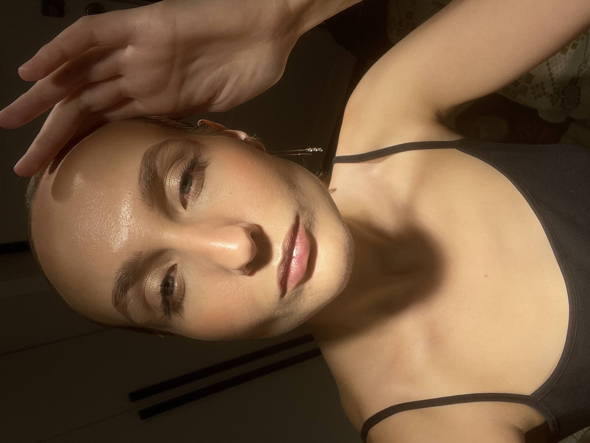 The image shows a close-up of a young woman's face and upper body, illuminated by warm, directional lighting that creates highlights and shadows. She has a bare head, with her hand resting on her forehead. Her eyes are partially closed, and her lips are slightly parted. She is wearing a black tank top, and a delicate earring with three small charms dangles from her right ear. The background is dark and out of focus, with vertical lines suggesting a wall or furniture. There are no specific details in the image that would allow for a determination of its location within the city of Chaikovsky, Russia.