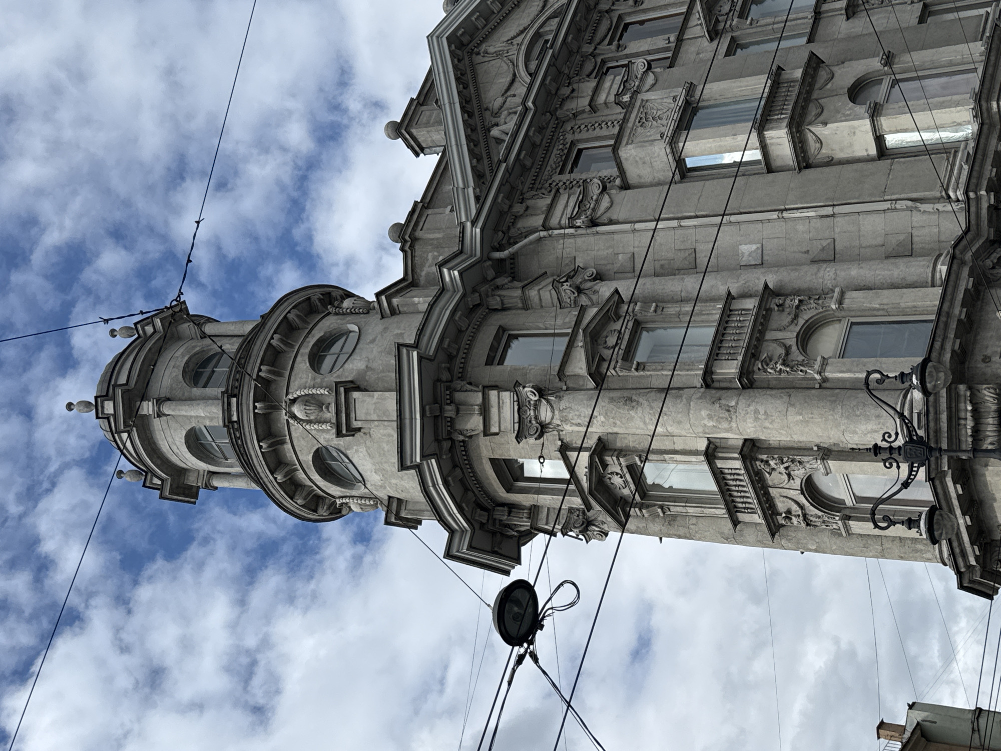 This image captures a detailed view of a corner of an ornate, historical building, likely from the late 19th or early 20th century, located in Saint Petersburg, Russia. The building features a prominent round tower with arched windows and decorative elements, including columns, cornices, and sculptures. The facade is constructed from a grey, weathered stone, indicative of the city's architectural style.
Several utility wires crisscross the frame, with a street lamp visible in the foreground, suggesting an urban setting. The sky is partly cloudy with blue patches, indicating daytime. The building's architecture, characterized by its classical influences and elaborate detailing, is typical of Saint Petersburg's historic center. There are no people visible in the image. The overall impression is one of grandeur and historical significance.