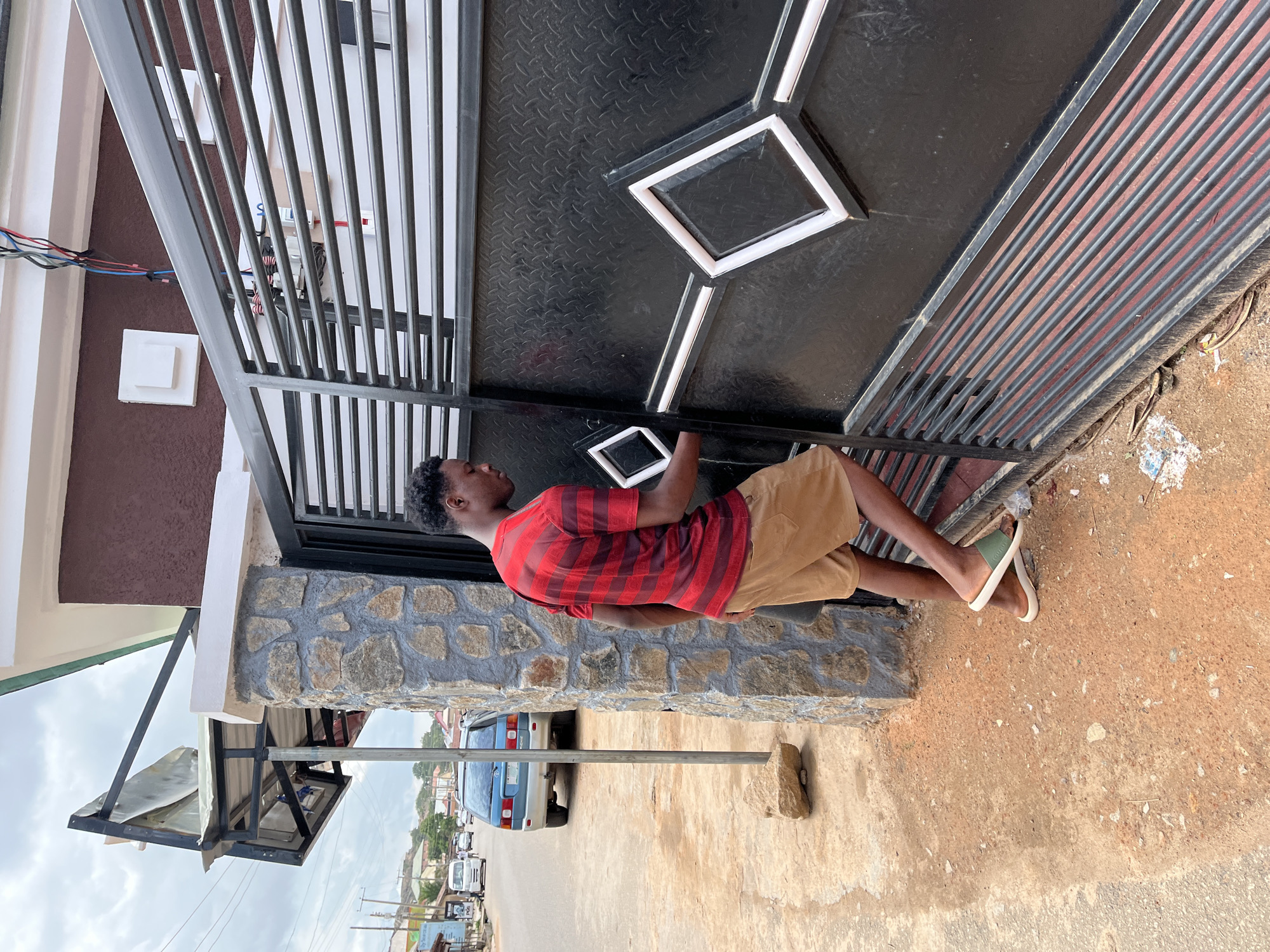 The image, taken in Jos, Nigeria, shows a young man standing beside a large black metal gate. He is positioned facing slightly to the right, holding a white tablet in both hands, appearing to be engaged with its screen. He wears a red and black horizontally striped short-sleeved shirt, light brown or khaki shorts, and green and cream open-toed sandals.
The gate, which forms a significant part of the scene, features a solid lower section with a distinctive raised diamond pattern accented by white frames, and an upper section composed of vertical black bars. To the man's left, a stone-clad wall serves as the gate's support, topped with a white border and a brown-painted section. Visible through the gate's bars, on the brown wall, are bundles of colored wires and some electrical components.
The man stands on unpaved, light brown ground. In the background, beyond the gate and wall, a street scene unfolds under an overcast sky, suggesting it is daytime. A light blue car is parked on the street, with other vehicles and buildings visible further in the distance. To the far left, a makeshift structure with a corrugated metal roof is visible. There is no clearly discernible text visible in the image.