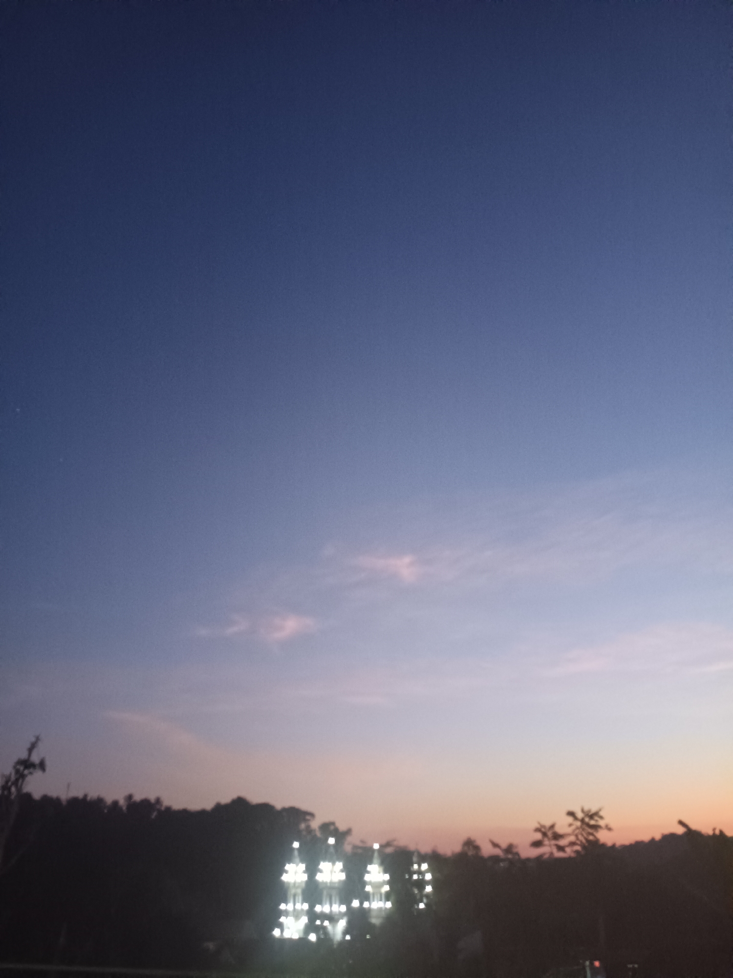 The image depicts a twilight scene with a gradient sky transitioning from deep blue at the top to soft purple and orange hues near the horizon. A silhouetted treeline runs across the lower part of the frame, interspersed with distant lights. On the right side of the treeline, there are several illuminated structures that appear to be buildings or perhaps decorative lights, casting a bright glow against the darkening landscape. The overall atmosphere is calm and serene, suggestive of an evening in Kadununggal, Indonesia. There are no people or animals visible in the frame.