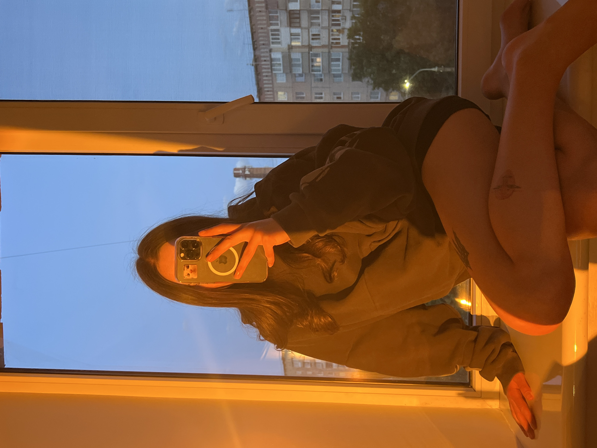 A woman is sitting on a windowsill indoors, taking a selfie with her smartphone. She is wearing a dark hoodie and black shorts. Her legs are crossed, and a small tattoo is visible on her right thigh. Through the window, a view of a multistory building is visible. The setting appears to be dusk or dawn. The location of the building visible through the window may be Ternopil, Ukraine.