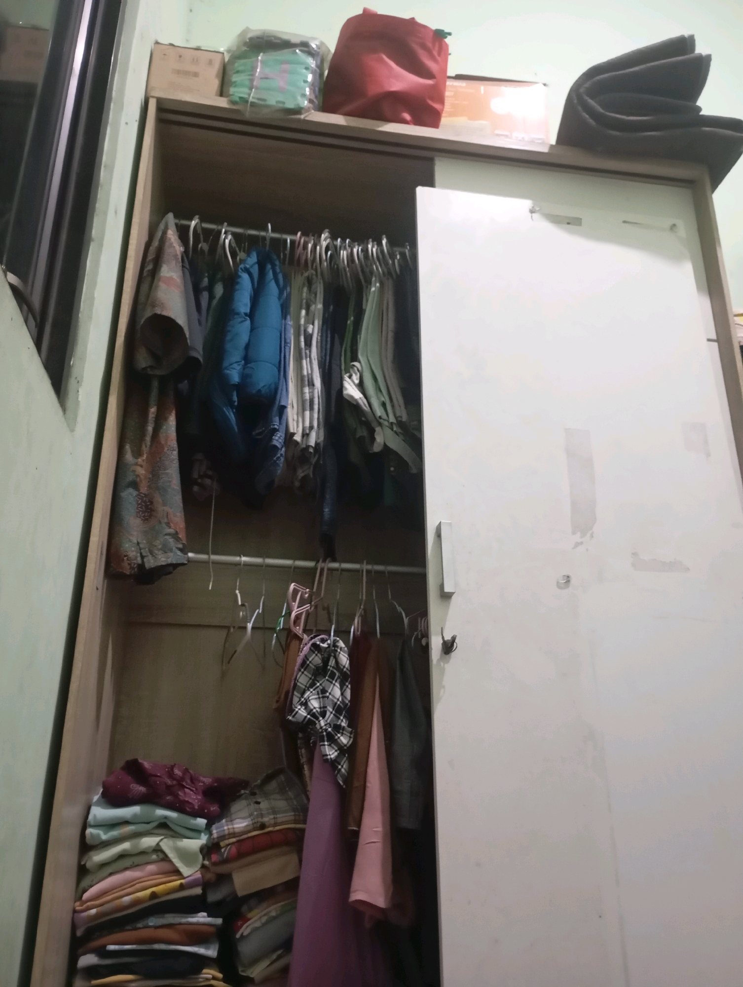The image shows the inside of a closet with its door ajar. The closet is filled with clothing. On the top shelf, there are several items including a red bag, a box, and a plastic-wrapped package containing green items. The main hanging section contains various garments on hangers, including what appears to be shirts and a blue jacket. The bottom section of the closet contains neatly folded stacks of clothing. The closet itself appears to be made of wood with a white sliding door. The environment suggests a domestic setting within a home.