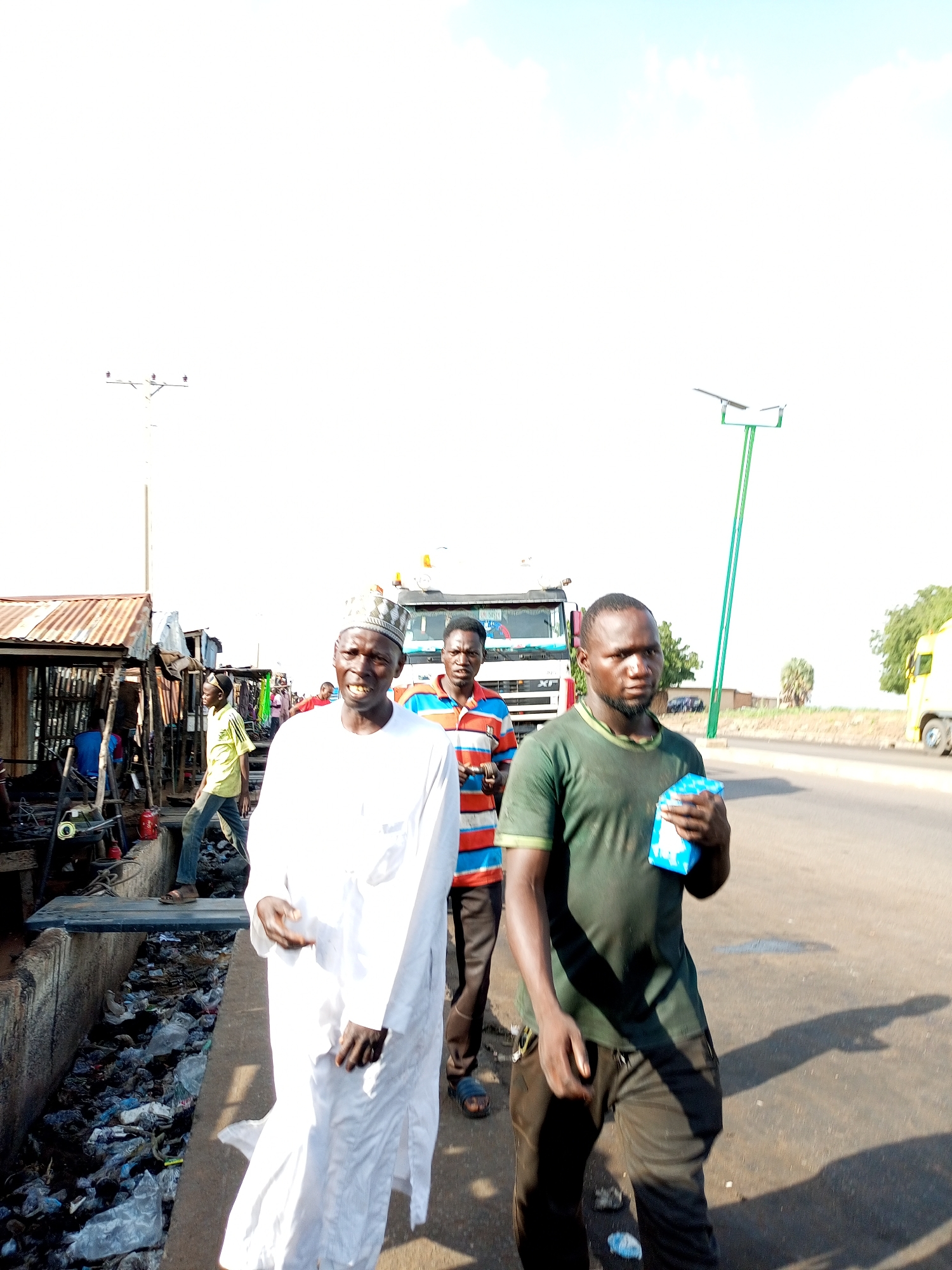 In Gadao, Nigeria, three men are captured in a photograph walking on a paved road. The man in the foreground, dressed in a dark green t-shirt and dark pants, is holding a small, light blue object, possibly a packet of tissues or wipes. He has a neutral expression and appears to be looking towards the camera. To his left, another man is walking, wearing a white traditional robe and cap, his gaze also directed forward. Behind them, a third man in a horizontally striped t-shirt is partially visible, his attention seemingly focused ahead.
The setting appears to be a roadside market or commercial area. To the left of the frame, there are makeshift stalls with corrugated iron roofs, suggesting a bustling, possibly informal, business environment. A large white truck is parked behind the individuals. A bright blue solar-powered street light stands tall on the right side of the road, indicating a more developed infrastructure alongside the market. The ground is littered with plastic waste, particularly along the edge of the road and in a drainage ditch.
The lighting suggests it is daytime, with the sun high in the sky, casting distinct shadows. The sky is a pale blue with some wispy clouds, indicating fair weather. The overall impression is a snapshot of daily life in a Nigerian community, highlighting the juxtaposition of commercial activity, everyday routines, and environmental challenges. There is no discernible text visible in the image.