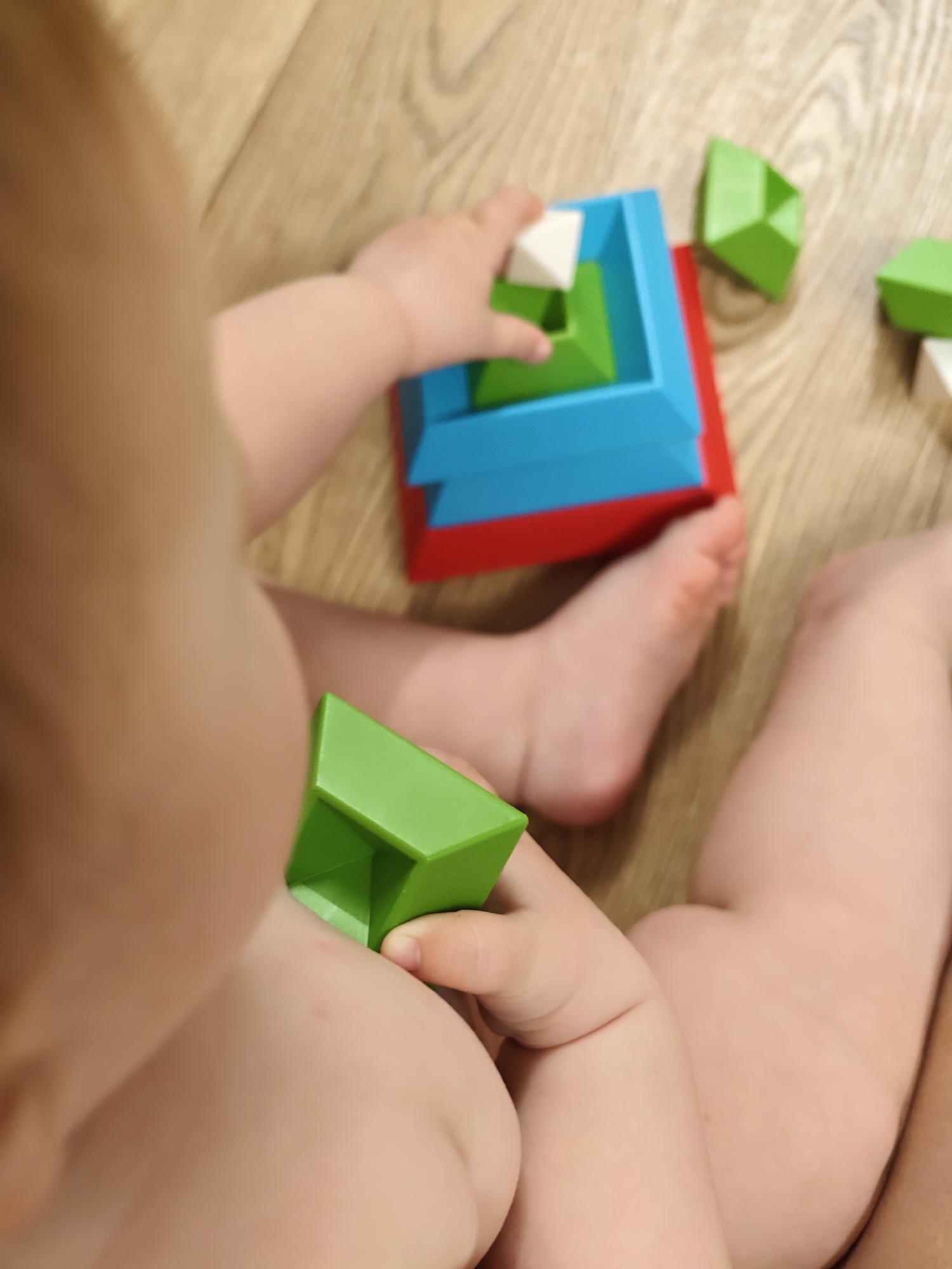 A young child is sitting on a light-colored wooden floor and playing with a set of colorful, geometric stacking blocks. The blocks are primarily red, blue, green, and white. The child is holding a green block in their left hand and appears to be placing a white block on top of a stack. The child is undressed. The location is not discernible as Chaikovsky, Russia, or any specific location.