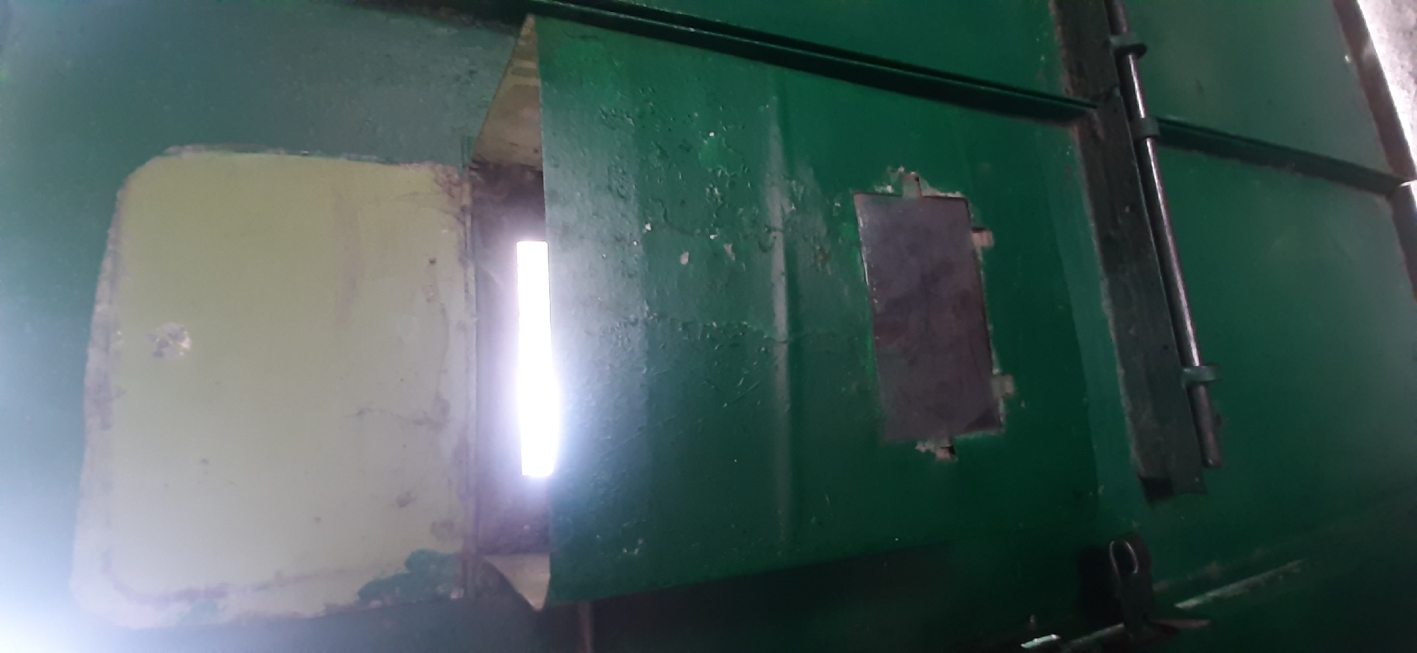 The image shows a close-up, vertical view of a green, industrial-looking object, likely a container or a piece of equipment. It is painted dark green, with areas of wear and peeling paint, revealing a lighter color underneath. The object features a rectangular opening at the top, with a lighter interior suggesting a light source from behind. Below this opening, there is a smaller, darker rectangular panel, possibly a viewing window or a label. The lower portion of the object displays metal hardware, including latches and rods. The overall impression is one of weathered, utilitarian construction. No specific location within Yessentukskaya, Russia is identifiable from the visual information provided.