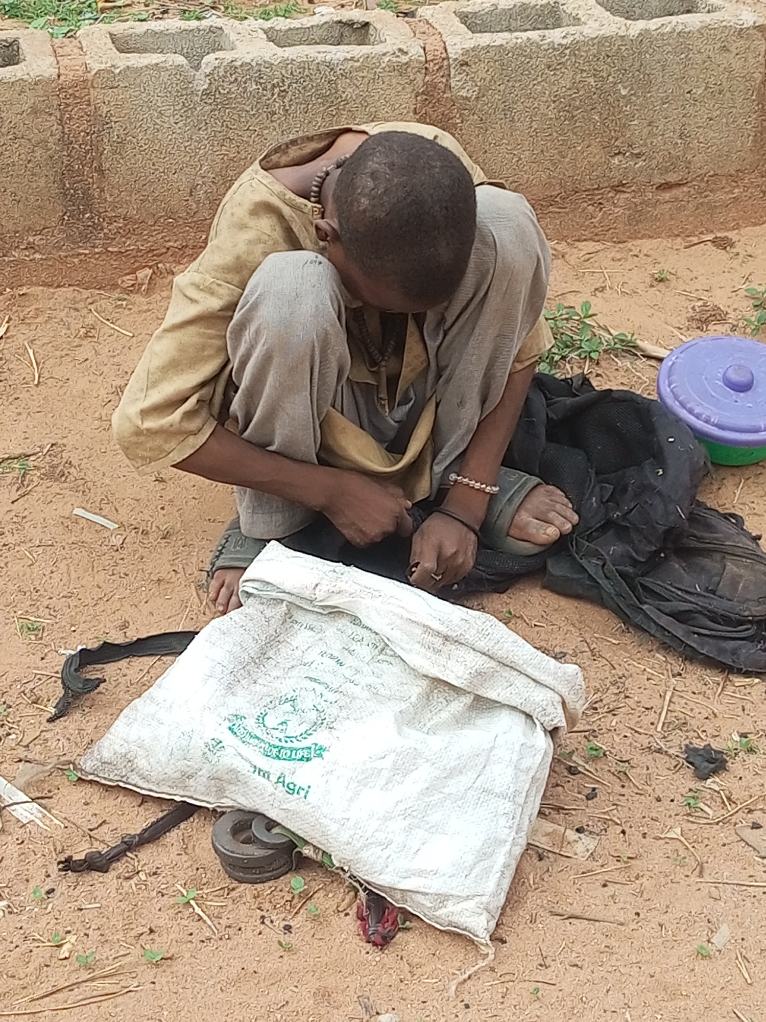 A person sits on the ground in an outdoor location. The ground is reddish-brown and appears to be soil or sand. A beige shirt and grey pants cover the person, who is sitting with their legs bent. A necklace is visible around the person's neck. The person's hands are in front of them, and they appear to be manipulating something. A white sack is in front of the person with some text on it. Beside the person is a black backpack and a purple and green plastic container. Behind the person is a low wall of what appears to be concrete blocks. There are no distinguishing landmarks to confirm the location as Tempure, Nigeria.