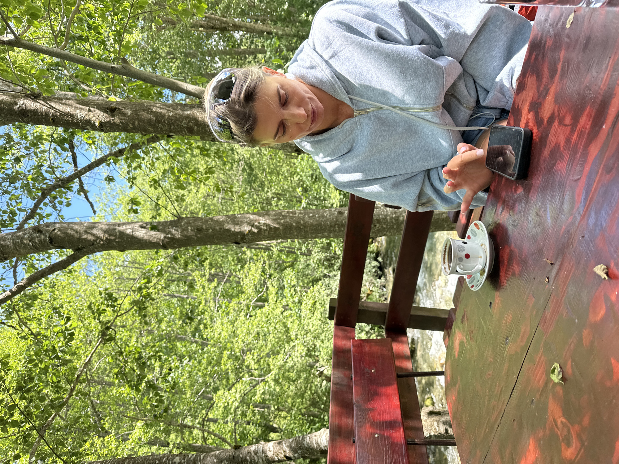 In the city of Bare Kraljske, Montenegro, a woman wearing a light blue hooded jacket and sunglasses is sitting at a red wooden table outdoors, with a stream visible in the background. She is looking down at her phone, which is placed on the table. Next to the phone is a white coffee cup with a decorative pattern and a saucer, containing a dark liquid. The table has a distressed finish with visible wood grain and some scattered leaves. Behind the table, a wooden bench painted red is visible, with a wooden railing separating it from the stream. The environment is lush and green, with numerous trees and foliage, indicating a natural, possibly forested, setting. The lighting suggests it is daytime, with sunlight filtering through the leaves.